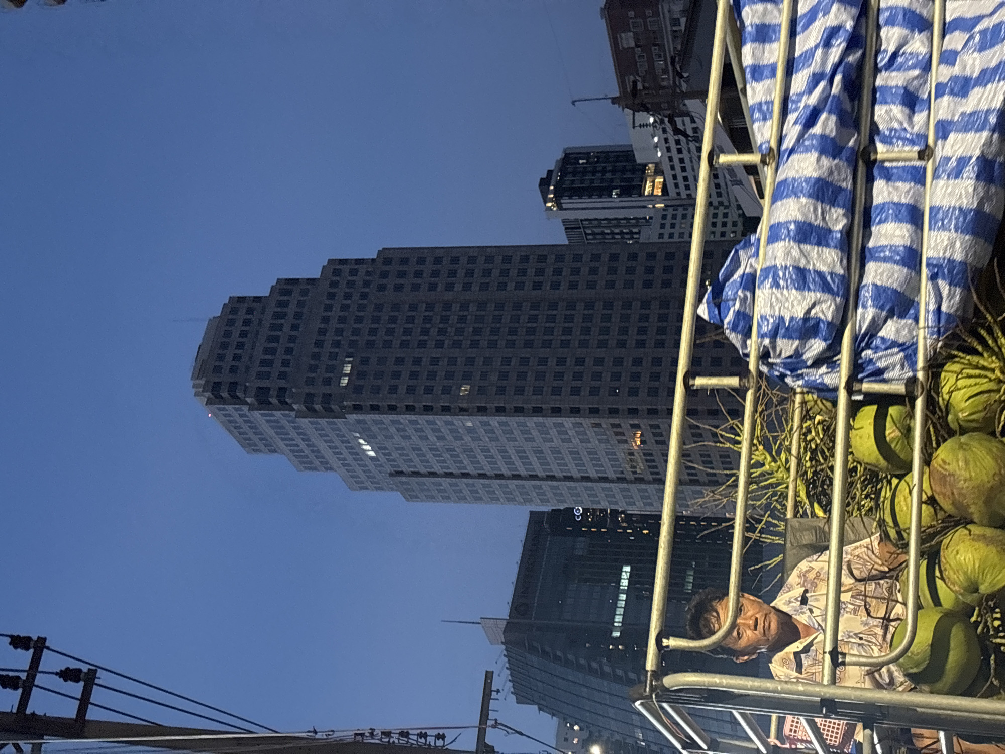 A low-angle evening shot captures a man in a patterned shirt resting among coconuts on a metal cart, with buildings and electrical lines in the background. The largest building is the Sathorn Square office tower in Bangkok, Thailand, due to its distinctive stepped design. The sky is dusky. A blue-and-white striped tarp covers part of the cart.