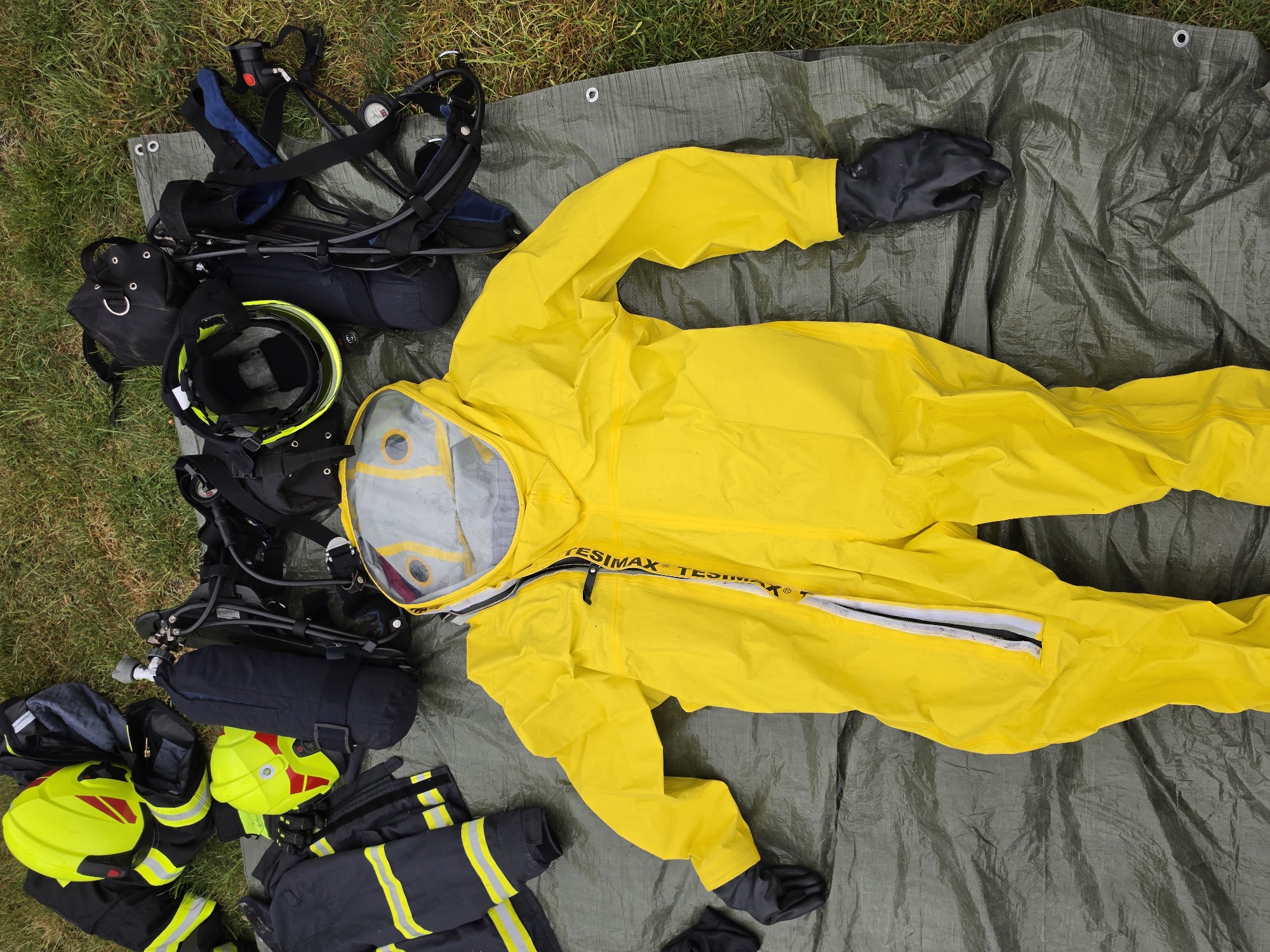 The image shows various pieces of emergency response equipment laid out on a gray tarp, possibly for inspection or inventory, against a backdrop of grass. The equipment includes a bright yellow hazmat suit with black gloves, a self-contained breathing apparatus (SCBA), fire helmets, and firefighter turnout gear. The scene is situated outdoors. The image was likely taken in Mlčechvosty, Czechia, as the location was provided.