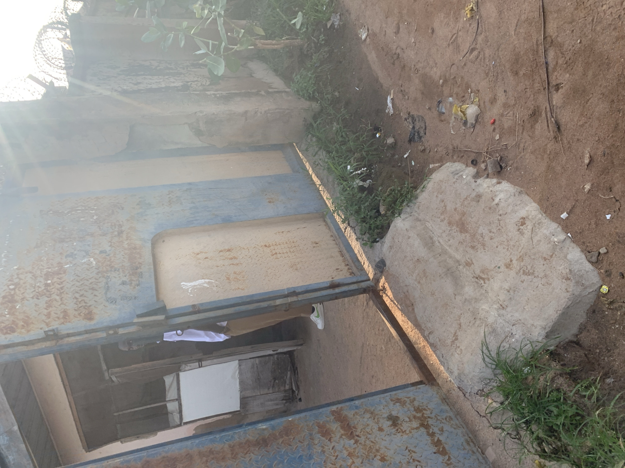 The image depicts an open, rusted metal gate leading into a structure. A person is visible in the doorway, wearing a white shirt and brown pants. The ground in front of the gate is dirt and scattered debris, with some vegetation growing near the edges. To the right of the gate, a concrete wall topped with barbed wire is visible, with a small tree growing beside it. The overall impression is one of a somewhat dilapidated or unkempt outdoor area.