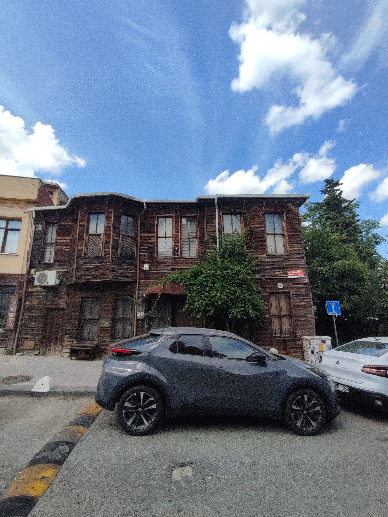 The image depicts a street scene in Kadıköy, Turkey, with an old wooden building and two parked cars. The building, likely a historic structure, is made of weathered wood and features multiple windows, some with intricate wooden shutters. An air conditioning unit is visible on the exterior wall. In front of the building, a dark grey hatchback is parked, angled towards the viewer. To its right, a white car, partially visible, is also parked. A speed bump is present in the foreground, indicating a residential or low-speed traffic area. A blue traffic sign with a white symbol is visible on the right side of the image, alongside a garbage bin covered in graffiti. Lush green trees grow behind the building, contrasting with the urban environment. The sky above is a vibrant blue, dotted with fluffy white clouds, suggesting a clear, sunny day. A street sign with Turkish text, "UZUHAFİZ", is affixed to the wooden building.