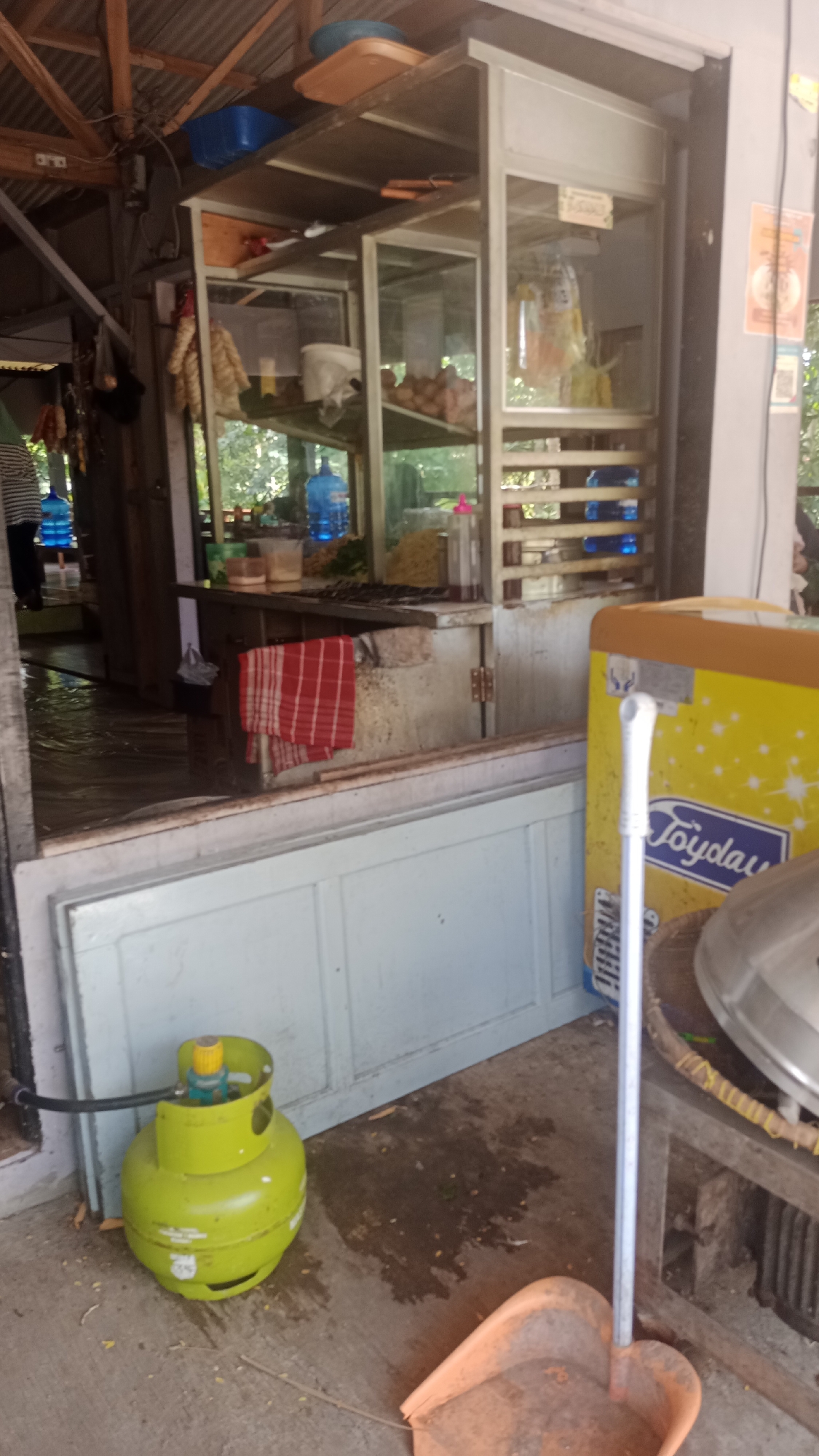 The image depicts an outdoor food stall in Cikalong, Indonesia. In the foreground, a bright green gas cylinder is placed on the concrete ground, connected by a black hose to the left. Next to it, an orange dustpan rests on the ground. To the right, a yellow and orange refrigerator with the "Joyday" logo is visible. Behind the refrigerator, a metal utensil with a white handle is standing upright. The stall itself features a glass display case showcasing food items. Various items, including bottles of water, are arranged on shelves within and around the stall. A red and white checkered cloth hangs on the side of the stall. The overall scene suggests a functional, everyday setting at a local food vendor.