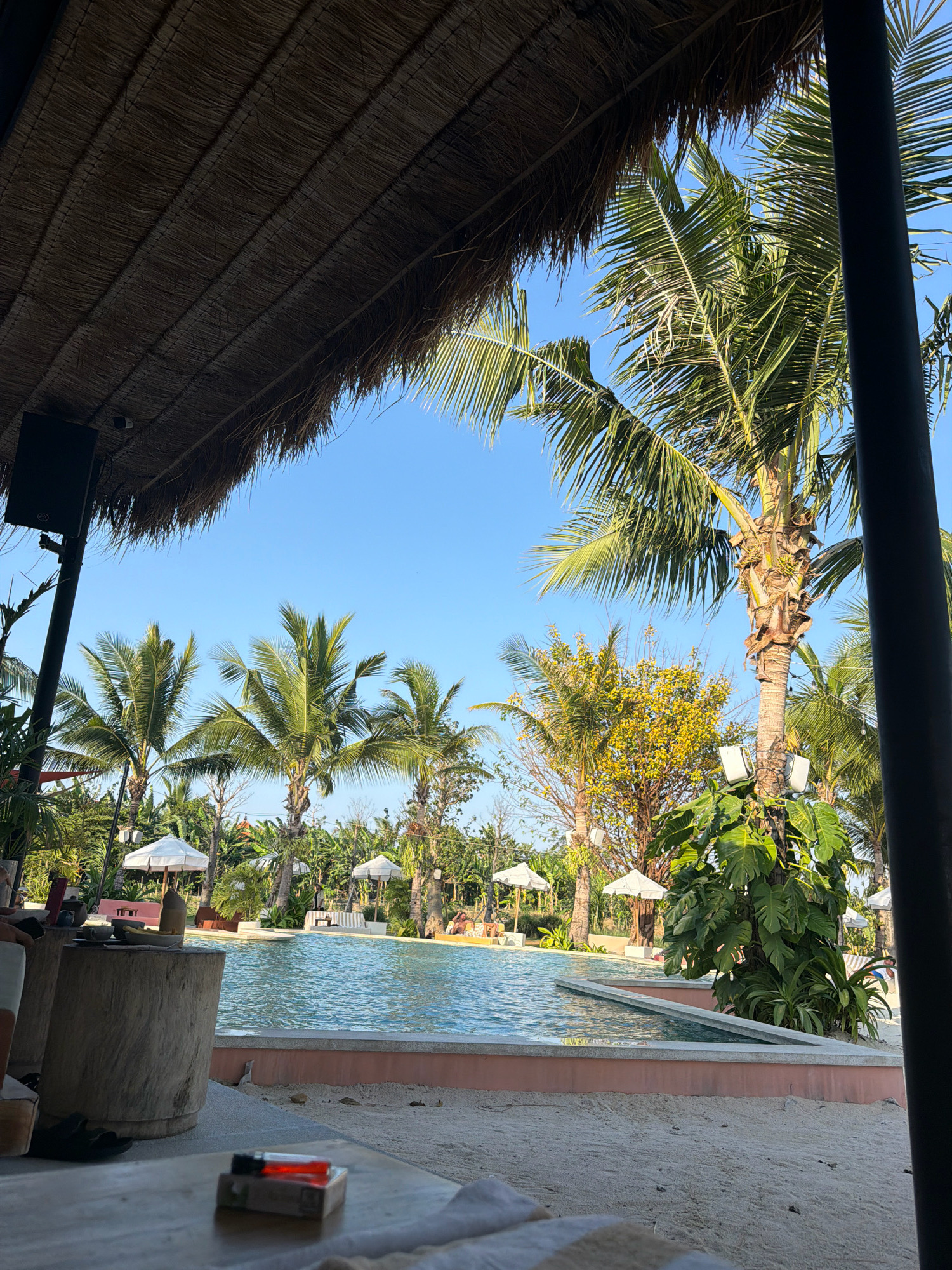 A poolside view is captured from a sheltered area with a thatched roof overhead. In the foreground, a wooden table holds a pack of cigarettes and a lighter with a lit flame. To the left, a large wooden stump serves as a side table, with a phone and other small items resting on it. Beyond the table is a swimming pool with clear blue water and a pink tiled edge. Several white umbrellas dot the area around the pool, offering shade. In the background, lush green palm trees and other tropical foliage dominate the landscape under a clear blue sky. The overall setting suggests a relaxing tropical resort or cafe. This scene is located in the city of Mengwi, Indonesia.