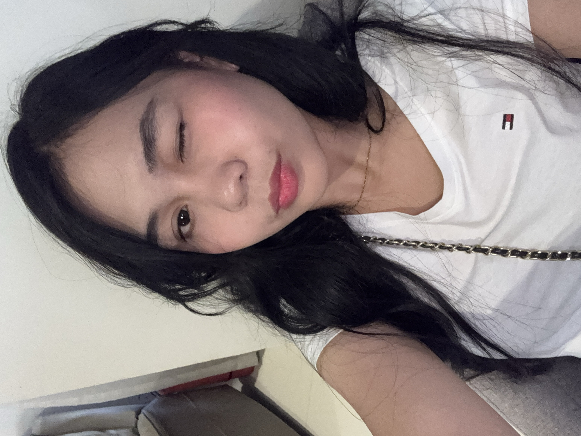 A young woman with long, dark hair is captured in a close-up selfie. She is winking and her head is slightly tilted. She is wearing a white t-shirt with a small Tommy Hilfiger logo on the left side of her chest. A delicate gold necklace is visible around her neck. Her makeup includes pink lipstick and blush. The background is a plain, light-colored wall, with a hint of furniture and a floor to the left of the frame. The overall lighting suggests an indoor setting. No specific location in Davao City can be identified from the visual information provided.