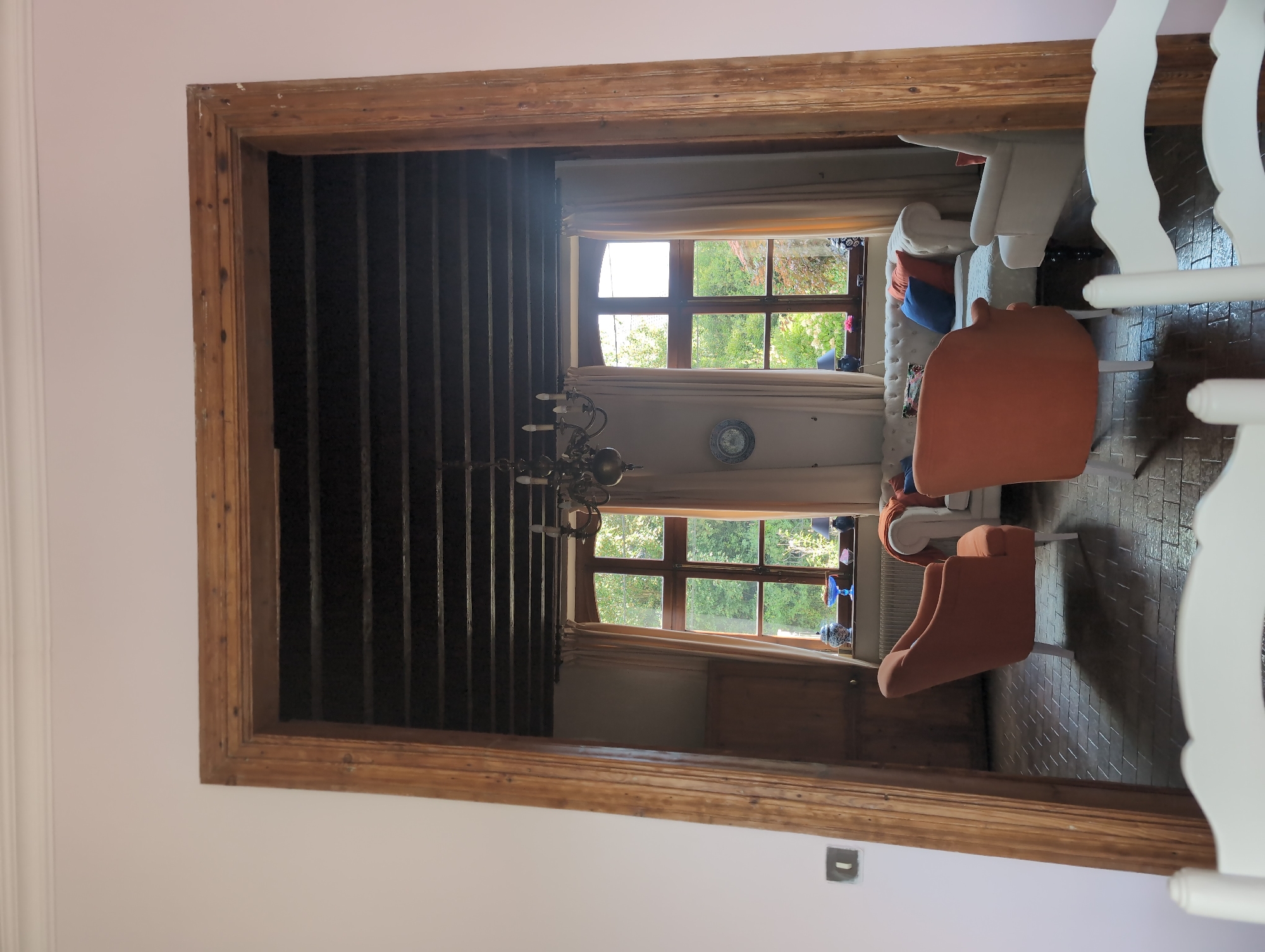This image depicts an interior view of a room through a wooden doorway in a building located in Contes, France. The room features a tufted white sofa with colorful throw pillows, two orange armchairs, and a white armchair. Two large windows with wooden frames are visible, draped with beige curtains, and overlook a green outdoor space. A chandelier hangs from the dark, exposed wooden beam ceiling. The floor is tiled in a diamond pattern. In the foreground, a portion of a white chair is visible. The lighting suggests it is daytime.