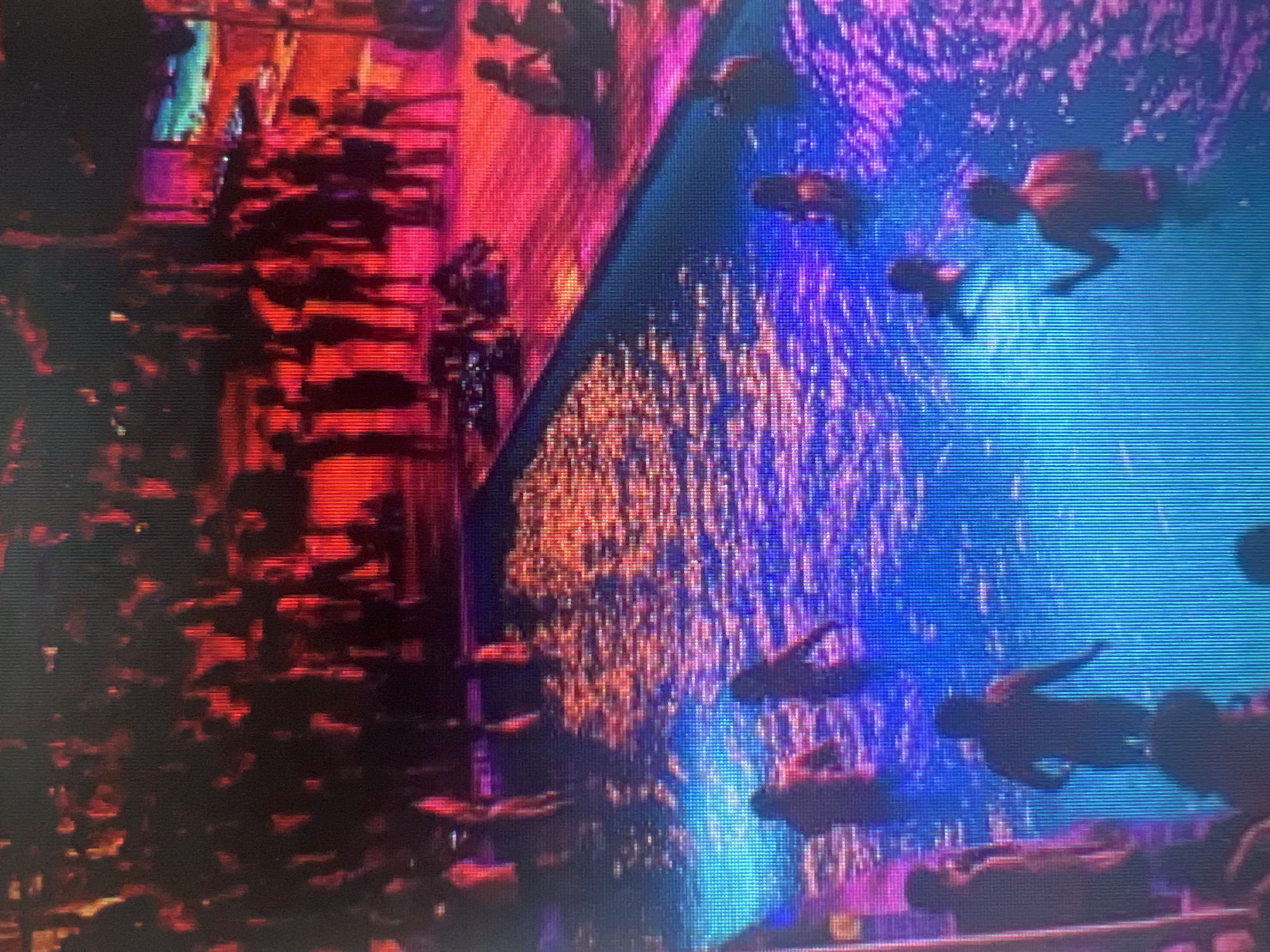 The image depicts a nighttime scene at an outdoor venue, possibly a club or party, in Cheb, Czechia. A group of people are gathered around and in a swimming pool illuminated with blue and pink lights. Some individuals are standing on a wooden deck area, while others are in the water. The lighting creates a vibrant, somewhat hazy atmosphere. The overall impression is one of a lively social gathering.