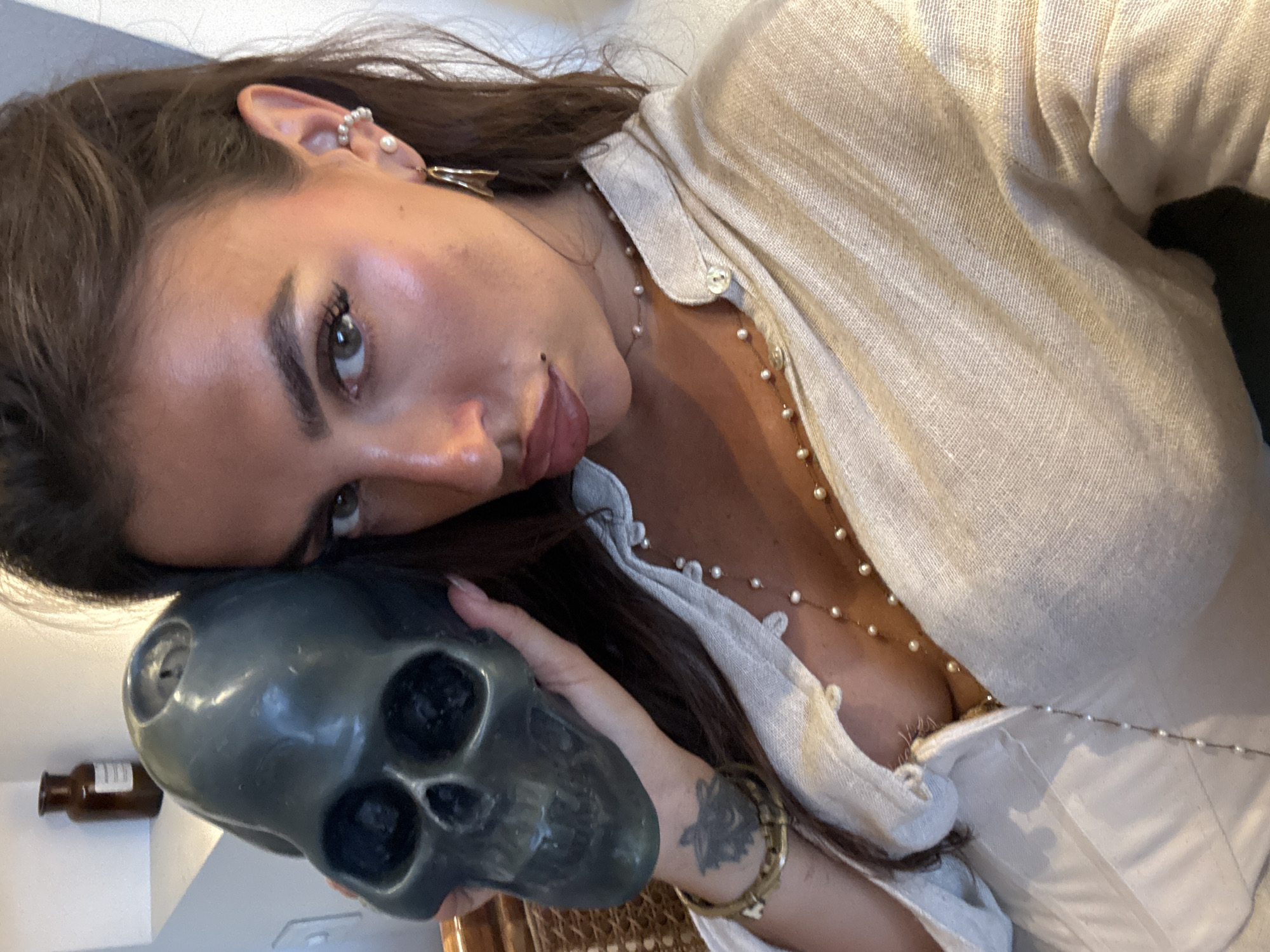 A woman with long, dark brown hair is posing for a selfie while holding a gray skull. She is wearing a light-colored, linen-like button-up shirt with pearl necklaces and earrings. A tattoo is visible on her wrist. A small amber glass bottle is on a white surface in the background. The location appears to be indoors, possibly a residence in North Kuta, Indonesia.