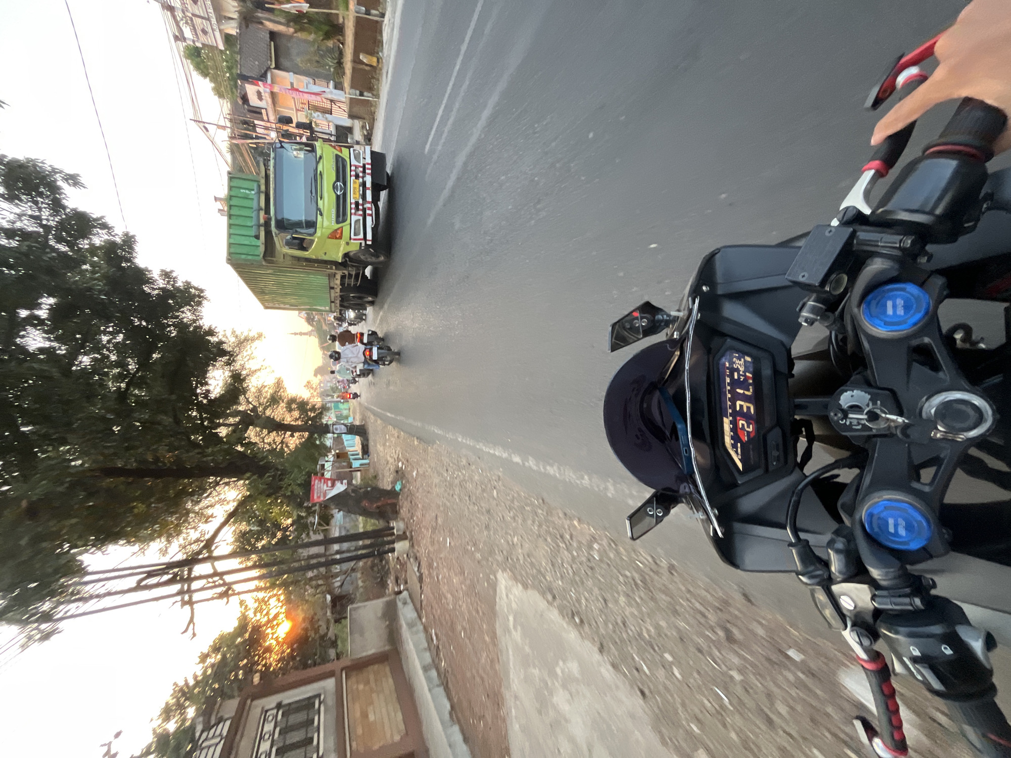 The image is a first-person perspective from a motorcycle on a road in Pelang, Indonesia, during sunset. The road is paved with asphalt and has visible lane markings. On the right side of the road, there are buildings, trees, and street lights. A large green truck is driving in the same direction as the motorcycle, and several other motorcycles are also on the road. The motorcycle's dashboard is visible in the foreground, displaying speed and other information. The sky is hazy with the sun low on the horizon, casting a warm glow on the scene.