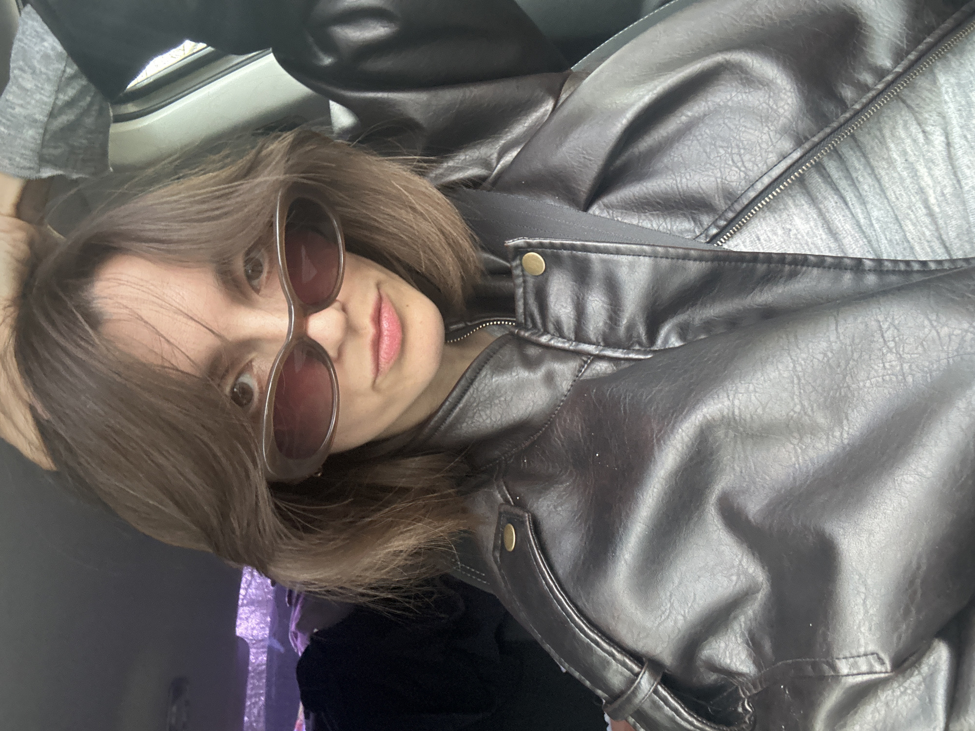The image is a close-up, tilted selfie featuring a young person, likely female, looking directly into the camera. She has medium brown, shoulder-length hair styled casually, partially obscuring her forehead. She wears oversized, round sunglasses with brown-tinted lenses. A hint of light pink lipstick is visible on her lips, suggesting a neutral to slightly smiling expression.
She is dressed in a dark brown or black faux leather jacket with a textured, slightly wrinkled appearance. The jacket features a prominent zipper closure down the front, a classic collar, and brass-colored snap buttons on the lapels. Beneath the jacket, a grey heathered t-shirt is visible.
The setting appears to be indoors, likely within a vehicle such as a car or train, given the visible window frame in the upper left corner. Through the window, blurred outdoor scenery and a faint purple light can be discerned. The lighting suggests it is daytime. The overall mood is casual. No text is visible in the image. The scene is contextualized as being in Moscow, Russia.