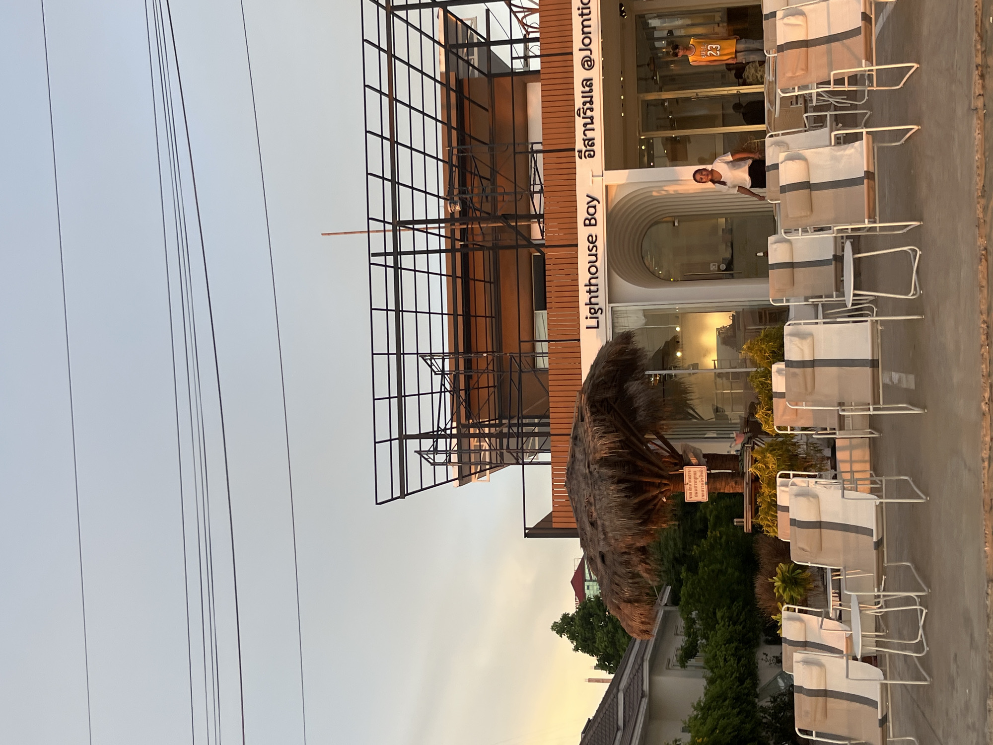 The image shows the exterior of a business called "Lighthouse Bay อีสานริมเล @Jomtic" in Pattaya City, Thailand. The building appears to be a restaurant or lounge, as suggested by the arrangement of outdoor seating with cushioned chairs. A partially obscured straw-roofed structure is visible in the foreground. A black metal frame structure is attached to the building's side. A sign near the straw structure reads in Thai, followed by "(ขอสงวนลิขสิทธิ)". Two people can be seen inside the building, one wearing a basketball jersey numbered "23". Power lines are visible in the sky above.