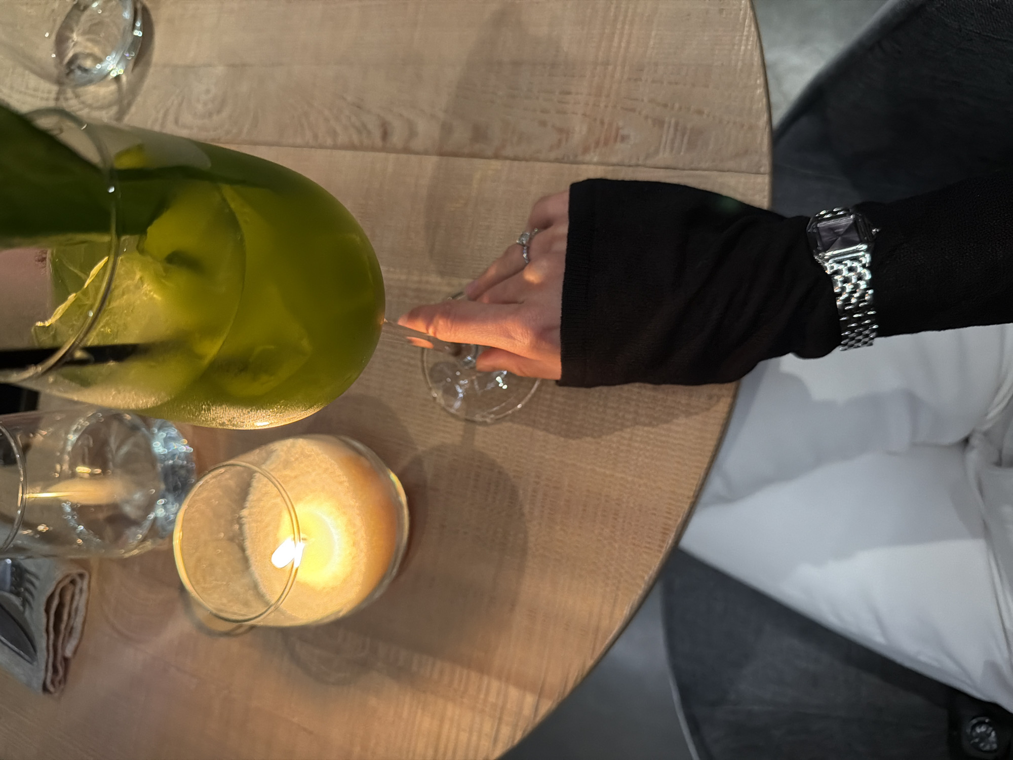 An overhead shot captures a cozy evening table setting, likely in a restaurant or bar in Krasnoyarsk, Russia. The scene is dominated by a light-colored wooden table surface with a subtle grain pattern.
In the foreground, a person's right arm and hand are visible, wearing a form-fitting black long-sleeved garment. On the wrist, a silver-toned watch with a rectangular dark face and a metal link bracelet is prominent. A sparkling ring with multiple stones adorns the ring finger of the hand. The hand is reaching for or resting near a clear wine glass on the table.
To the left, a large, clear glass pitcher filled with a vibrant green liquid, possibly a cocktail or infused drink with visible ice or fruit pieces, is positioned. Another empty clear glass is near the pitcher.
A central element is a lit candle encased in a glass holder, casting a warm, soft yellow-orange glow on the table and surrounding objects, suggesting a dim, intimate atmosphere. The flame of the candle is clearly visible. Further to the left, a folded grey or light brown cloth, possibly a napkin, is partially visible.
In the background, a white cushioned seat or chair is visible, along with darker furniture elements, reinforcing the indoor dining setting. The overall lighting is low and ambient, primarily from the candle. There is no visible text in the image.