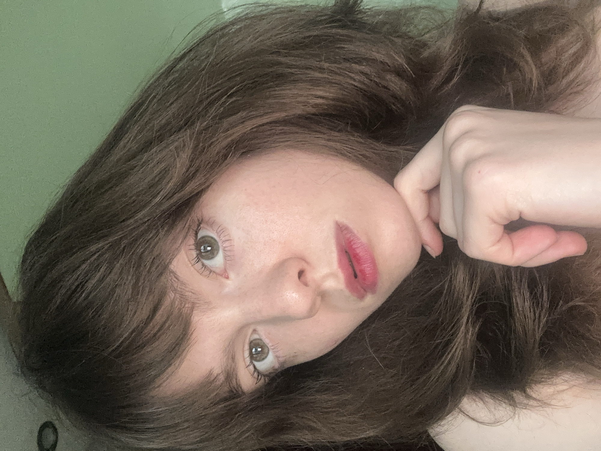 A rotated close-up portrait features a young adult female, likely taken indoors in Moscow, Russia. Her head and part of her upper body are visible, framed by her long, wavy dark brown hair that spreads across the foreground and background. Her fair skin features subtle freckles, and her light brown eyes look directly towards the camera, accentuated by visible eyelashes. Her lips are painted with a reddish-pink lipstick, appearing slightly diffused or uneven. One of her hands is visible near her face, with fingers gently curled. The background consists of a plain, light green wall. The subject's expression is calm and direct. There is no visible text or specific time-of-day indicators within the frame.