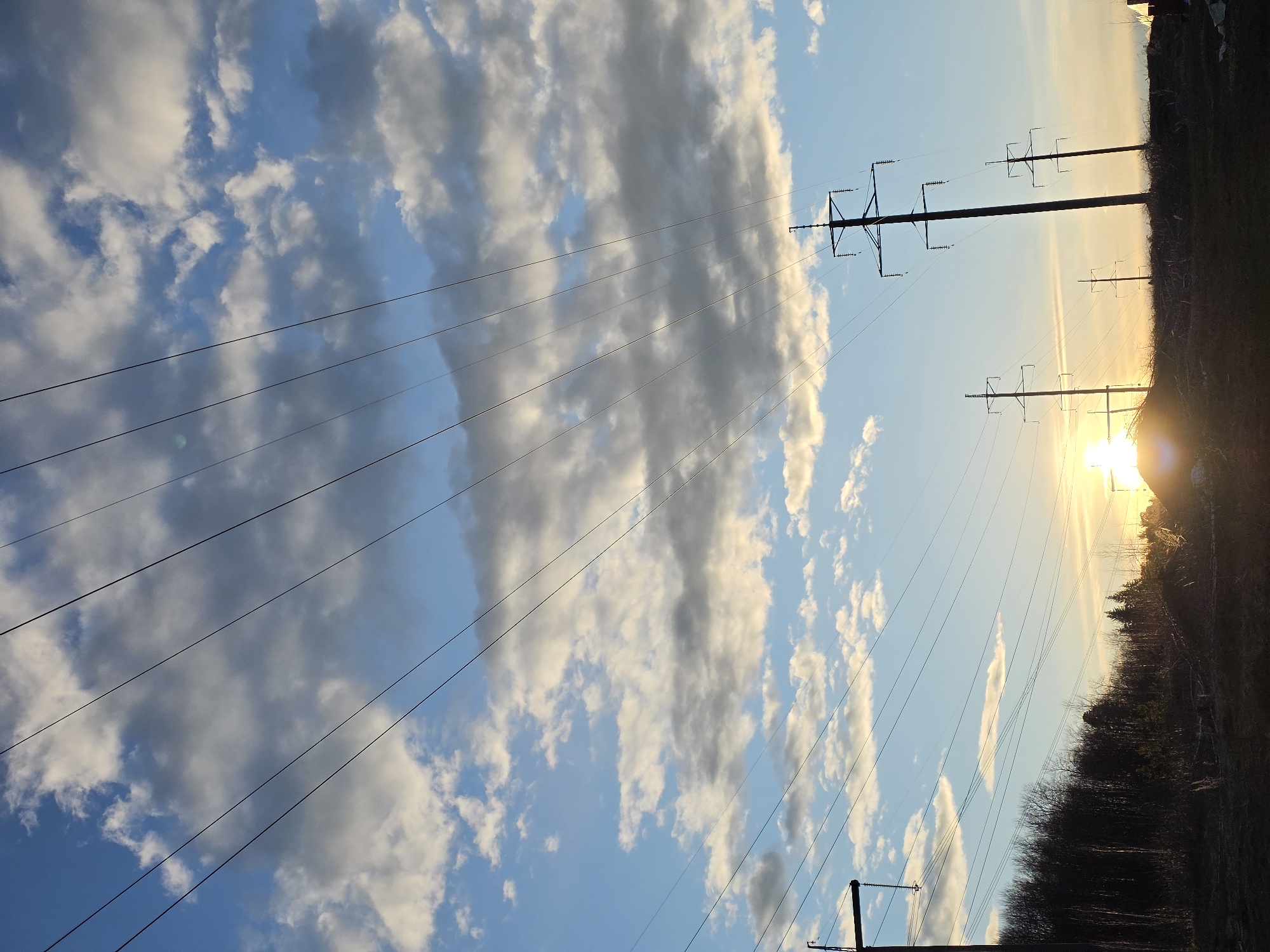 This is an outdoor wide shot featuring a vast sky, power lines, utility poles, and a sun low on the horizon. The sky is predominantly blue, filled with scattered white and grey cumulus clouds that vary in density and shape. Several dark, thin power lines stretch diagonally across the upper and mid-portions of the frame.
On the right side of the image, multiple dark utility poles or transmission towers stand tall, supporting the network of power lines. The sun is a brilliant, intense light source located low on the far right horizon, casting a bright glare and a warm, golden glow across that section of the sky. This strong illumination suggests either sunrise or sunset.
Along the bottom horizon, a dark, silhouetted landmass or treeline is visible, appearing to be dense foliage or a forest, contrasting sharply with the bright sky above. No specific human activity is taking place, and there are no discernible people or visible text. The setting appears to be a rural or semi-rural landscape, characterized by open skies and utility infrastructure. The weather is partly cloudy.