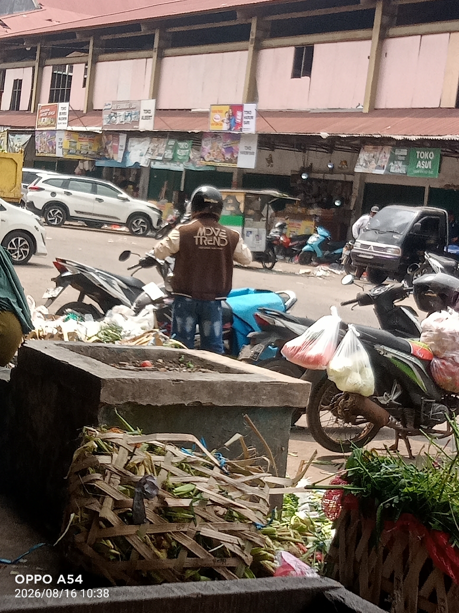 In Jambi, Indonesia, a street scene unfolds near a market. In the foreground, a weathered concrete structure holds a woven basket filled with dried vegetation. To the right, another woven basket is filled with fresh green leafy vegetables and some red produce. A motorcycle, its rear wheel prominent, is parked beside the concrete structure, with plastic bags filled with red and white produce hanging from it. In the mid-ground, a person wearing a brown jacket with "MOVE TREND" printed on the back and blue jeans is standing near a motorcycle, their back to the camera. Another motorcycle, with a blue seat, is parked further back. A black utility vehicle is visible in the background. Above, the facade of a building with multiple windows and advertising banners for various shops, including "TOKO ASUI," lines the street. The overall environment suggests a bustling market area with a mix of retail activity and pedestrian/vehicle traffic.