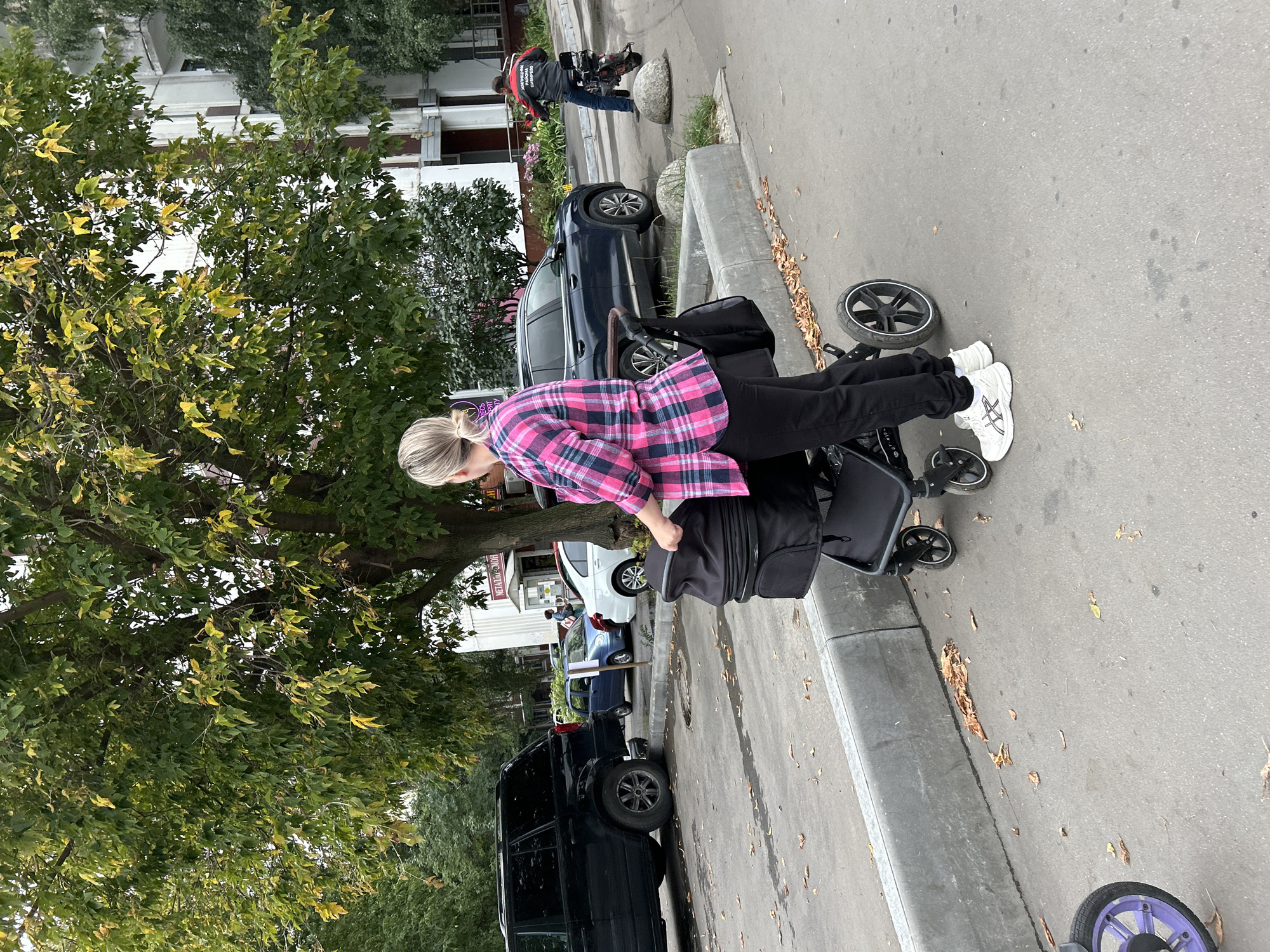 In Moscow, Russia, a woman with blonde hair pulled back in a ponytail, wearing a pink and blue plaid shirt and black pants, is pushing a black stroller. The stroller has a bag attached to the handle. She is wearing white athletic shoes with black accents. In the background, a dark SUV is parked to the left. Further to the right, a dark blue sedan is parked. A person on a moped, wearing a red and black jacket, is visible in the distance on the right side of the image. The scene appears to be an urban environment with a sidewalk and street. Trees with green and yellowing leaves are present, suggesting it is autumn. Buildings are visible in the background. Fallen leaves are scattered on the ground.