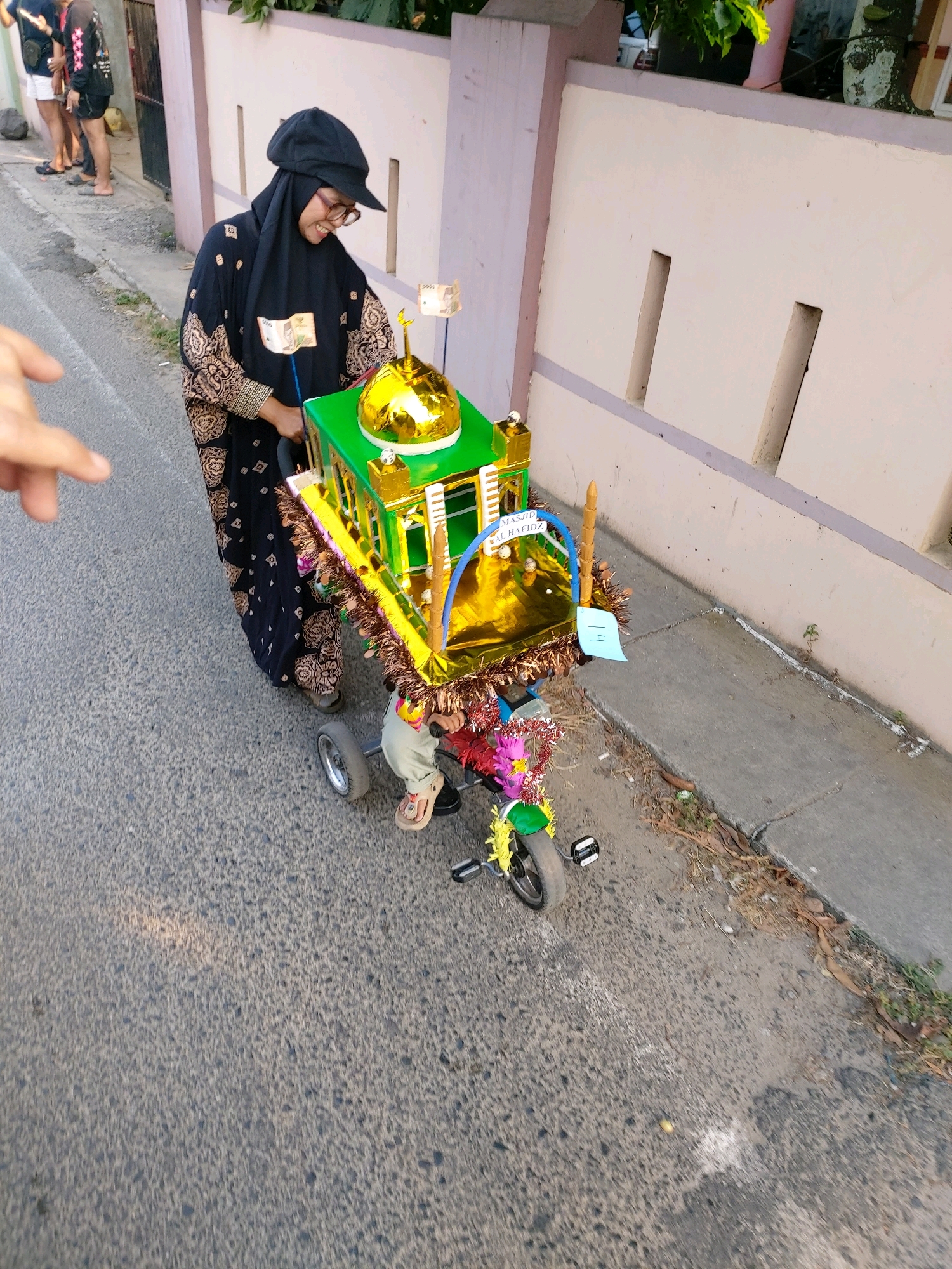A woman wearing a black hijab and patterned abaya pushes a decorated tricycle. The tricycle is adorned with a miniature mosque model, featuring a gold dome and green walls, and is decorated with colorful streamers and what appear to be paper bills. A child in a light-colored outfit sits on the tricycle. They are on a paved road next to a light-colored wall. The scene appears to be in Bandar Lampung, Indonesia, based on its architecture and the context of the tricycle decoration, which likely relates to a religious celebration.