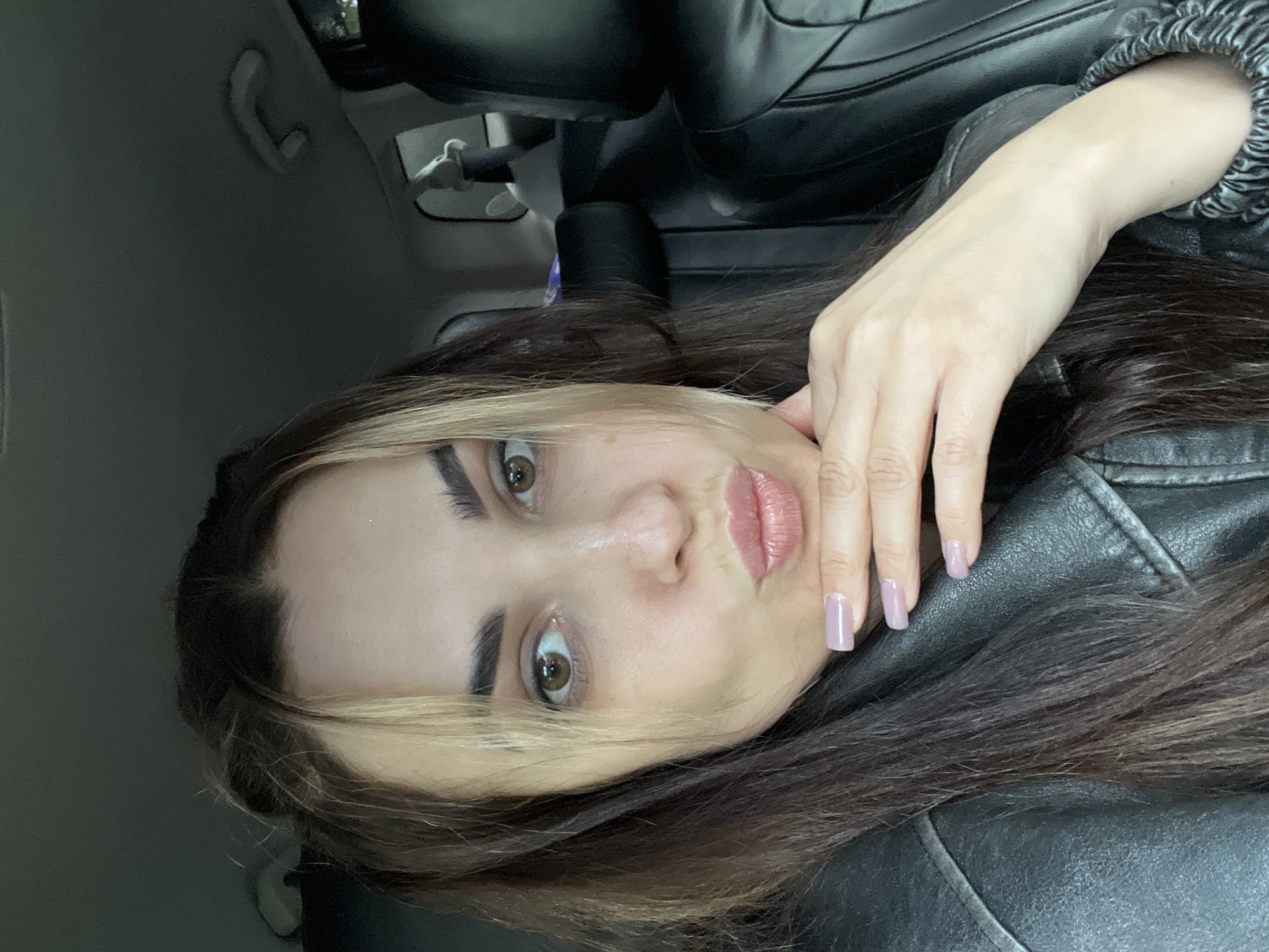 A close-up portrait of a young woman, rotated 90 degrees counter-clockwise, shows her looking directly at the camera from inside a vehicle. She has long, dark brown hair with distinctive blonde or light brown streaks framing her face. Her eyes are brown, and her eyebrows are dark and well-groomed. Her lips are full and appear to have a glossy pink or light red color. Her complexion is fair to medium.
One of her hands is gently posed near her face, with her manicured fingernails, painted a light purple or lilac color, lightly touching her chin and cheek. She is wearing a black jacket, which appears to be made of leather or a similar material, with some textured or ruched detailing visible on the cuff of the sleeve.
The setting is clearly the interior of a car. Visible background elements include a dark gray car headliner (ceiling) with a grab handle, dark car seats behind her, and a reflection of what might be a rearview mirror assembly in the upper right. The lighting appears to be natural daylight, softly illuminating her face. Her expression is neutral and composed, reflecting a posed moment. There is no visible text in the image.