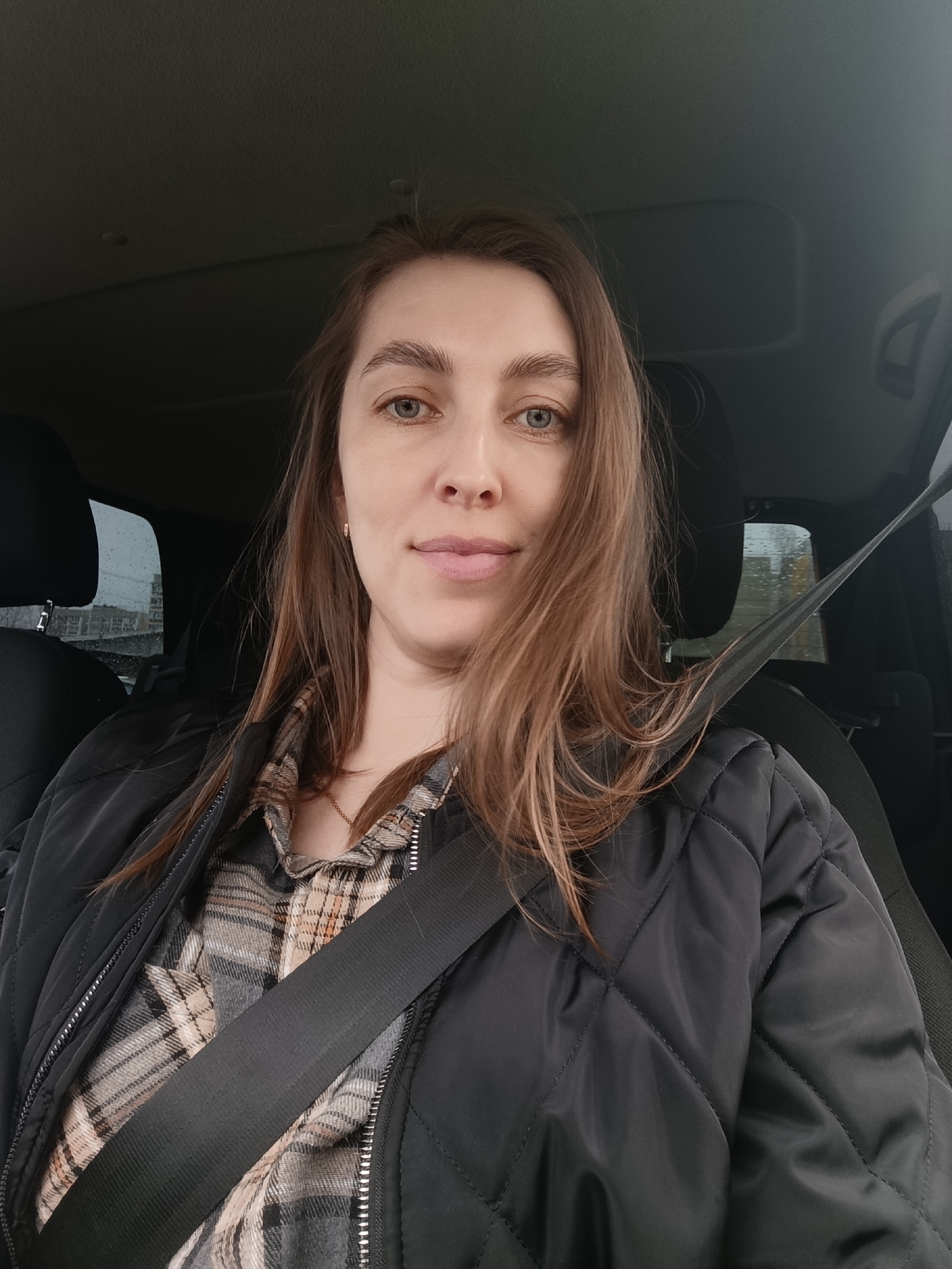 The image shows a woman in the driver's seat of a car. She is wearing a black quilted jacket over a plaid shirt and has a seatbelt fastened across her chest. Her hair is light brown and falls to her shoulders. Her eyes are blue. The background visible through the car windows appears to be a city scene, possibly Chaikovsky, Russia, although without additional information, location verification is impossible. The lighting suggests it is daytime. There is no interaction visible.