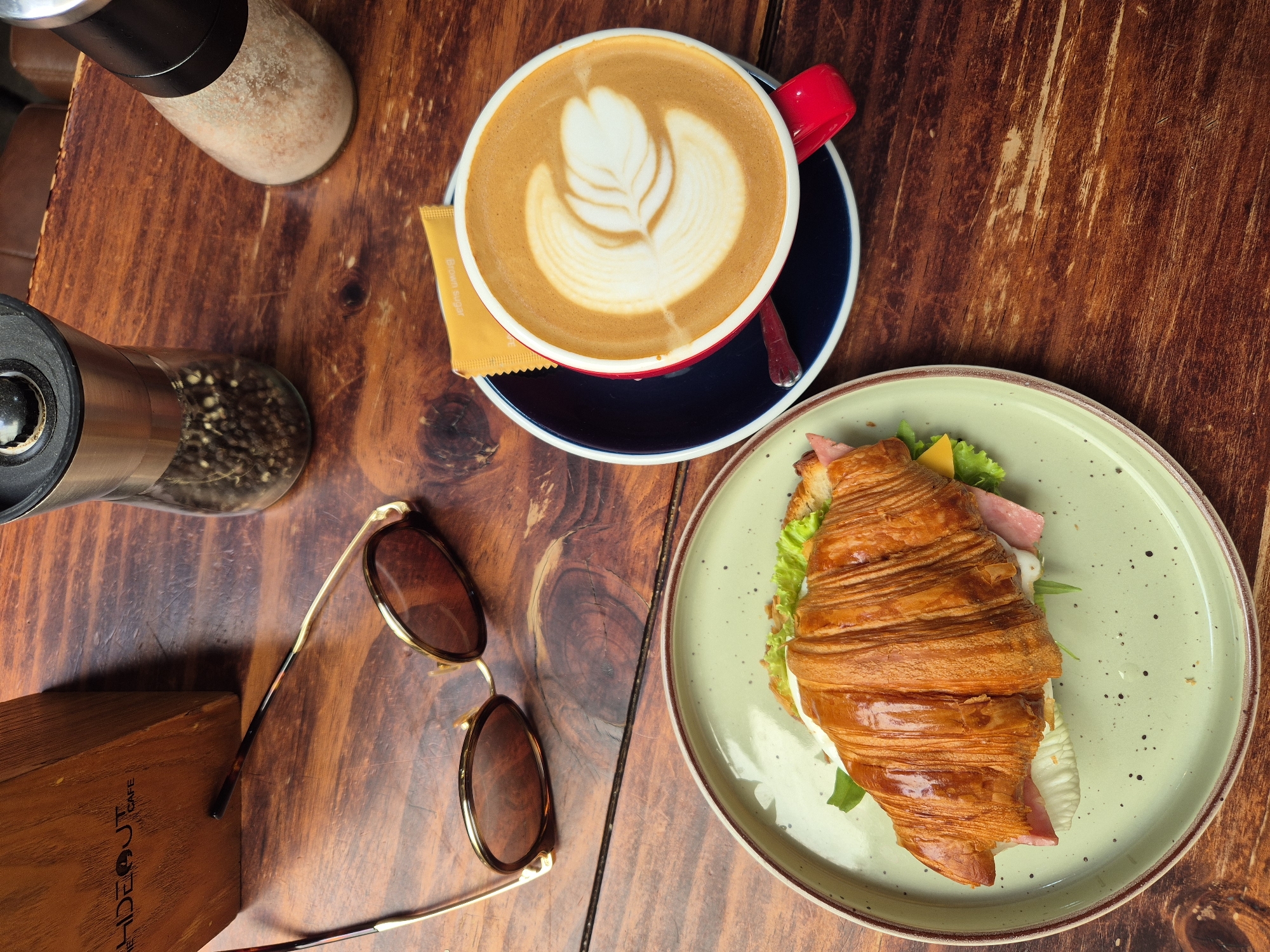 This is a top-down, eye-level, medium shot of a breakfast spread on a dark wooden table at "Hideout Cafe" in Da Nang, Vietnam. The cafe's logo is visible on a wooden sign in the upper left corner. In the foreground, a light green speckled plate holds a croissant sandwich filled with ham, cheese, and lettuce. To the right of the sandwich, a red coffee cup with a latte art design sits on a dark blue saucer. A packet of brown sugar is placed behind the coffee cup. In the background, a pepper grinder and a salt shaker are visible. A pair of gold-rimmed sunglasses with brown lenses rests on the table between the sign and the coffee. The lighting is natural and diffused, casting soft shadows.