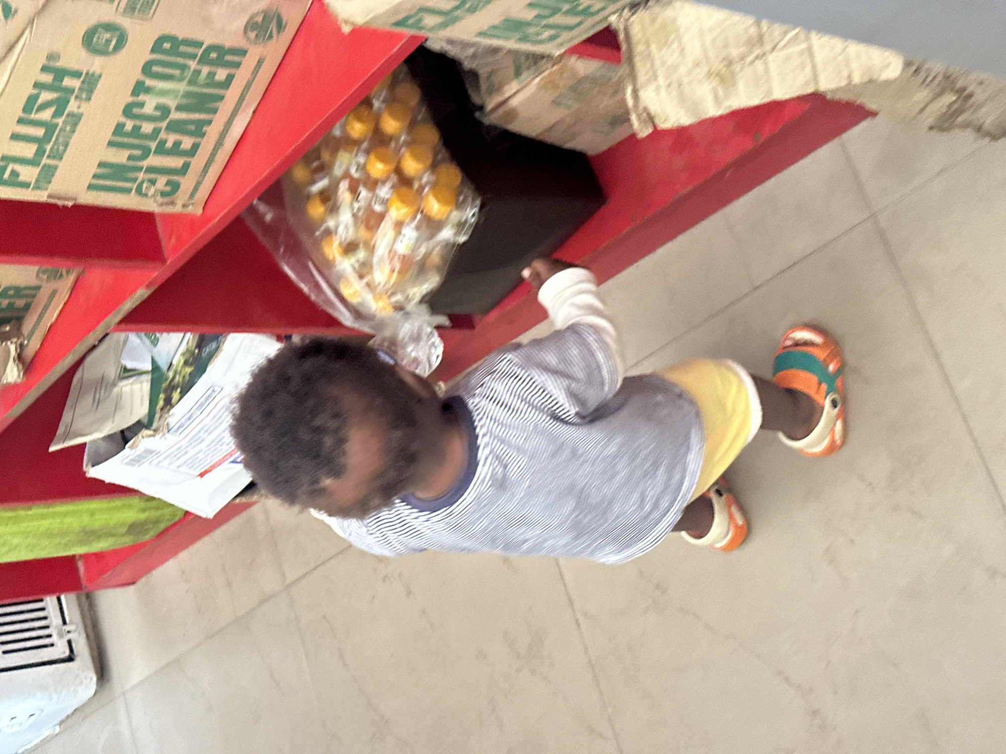 A young child, seen from above and from the side, stands in front of a red shelf. The child is wearing a blue and white striped t-shirt and yellow shorts, with orange and teal sandals on their feet. The child is reaching towards a clear plastic bag filled with small, yellow-capped bottles. The shelf is stocked with boxes, one of which has visible text: "FLUSH INJECTOR CLEANER". The floor is tiled with light-colored square tiles. The scene appears to be indoors, possibly a shop or market stall, given the shelving and products. The lighting suggests daytime. The overall impression is of a child interacting with items on a shelf.