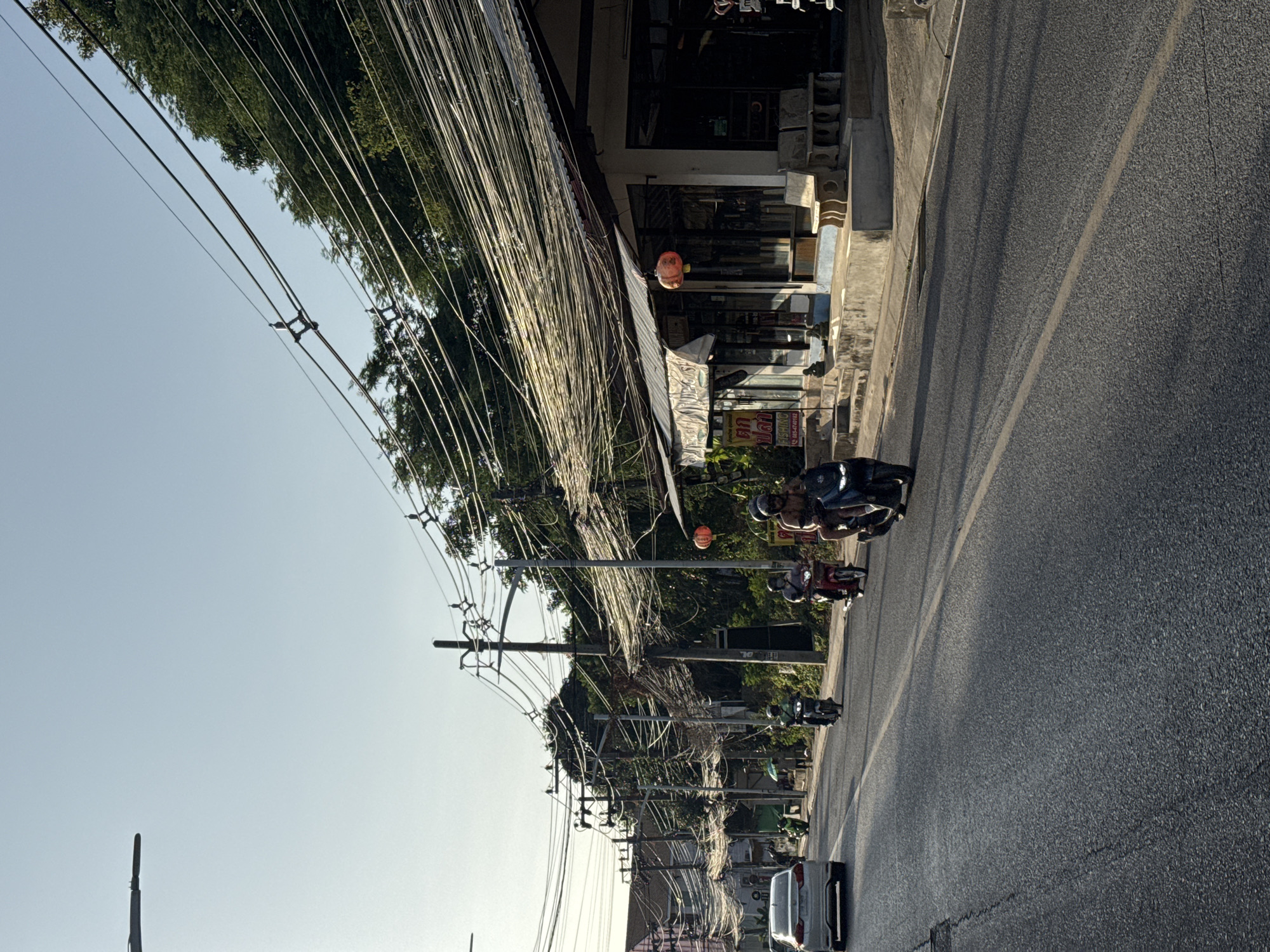 The image depicts a street scene in a city on the island of Ko Samui, Thailand. The road is paved and appears to be a two-lane street with traffic. Several motorcycles are visible, with riders wearing helmets, indicating they are actively being ridden. A white car is also present on the road. Along the side of the road, there are buildings with storefronts, and a prominent feature is the dense network of overhead electrical wires supported by numerous poles. Trees and vegetation are visible behind the buildings, suggesting a tropical environment. The sky is clear and bright, indicating daytime. Some shops have signs with local script. The overall impression is of a bustling street in a Southeast Asian city.