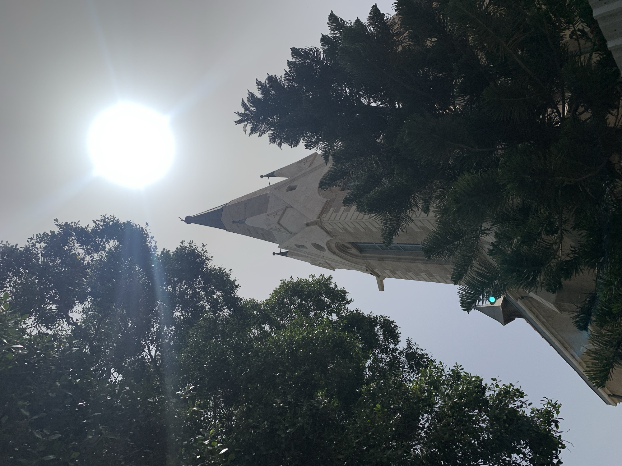 A low-angle shot captures a church steeple, partially obscured by trees, set against a bright sky with the sun prominently positioned in the upper left. The steeple, possibly of a historic structure, is of a light stone color, featuring gothic architectural details. Trees with dense foliage, both deciduous and evergreen, frame the lower and right portions of the composition. The sky is a pale, washed-out blue due to the sun's glare. The location is likely in Tel Aviv, Israel, based on the architecture and vegetation types common to the region.