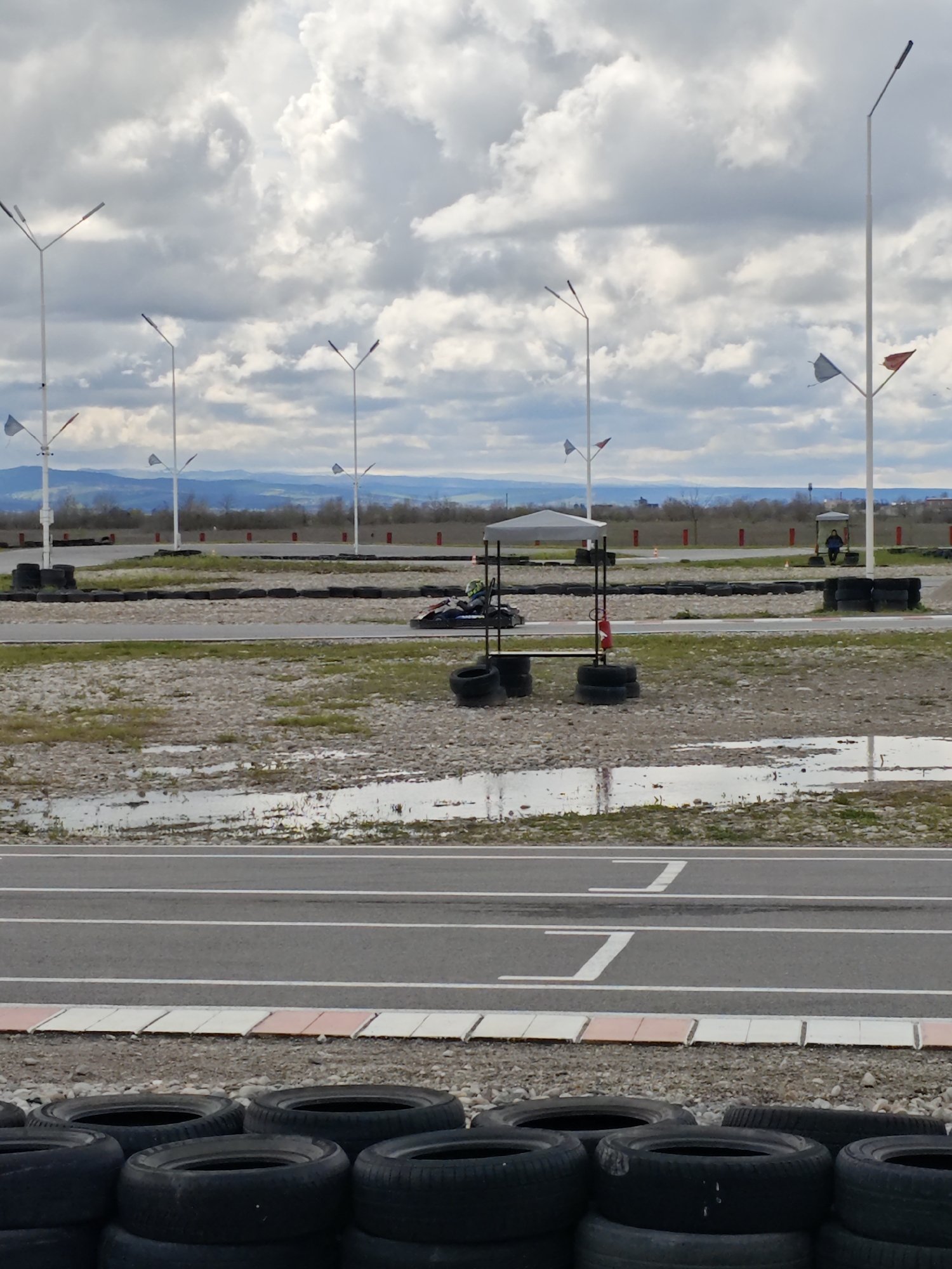 The image depicts a go-kart racing track in an unspecified area, potentially Kishpek, Russia. In the foreground are stacked black tires acting as a barrier. Behind the tires is a gray asphalt track marked with white lines, including the start/finish line. Further back, the track area is surrounded by gravel and patches of green vegetation, with puddles of water visible. Additional tire barriers line the track. In the distance, light poles stand tall, and a hilly landscape is visible under a cloudy sky. A go-kart is on the track, and a person is standing under a shelter. A red fire extinguisher is attached to a structure with tires.