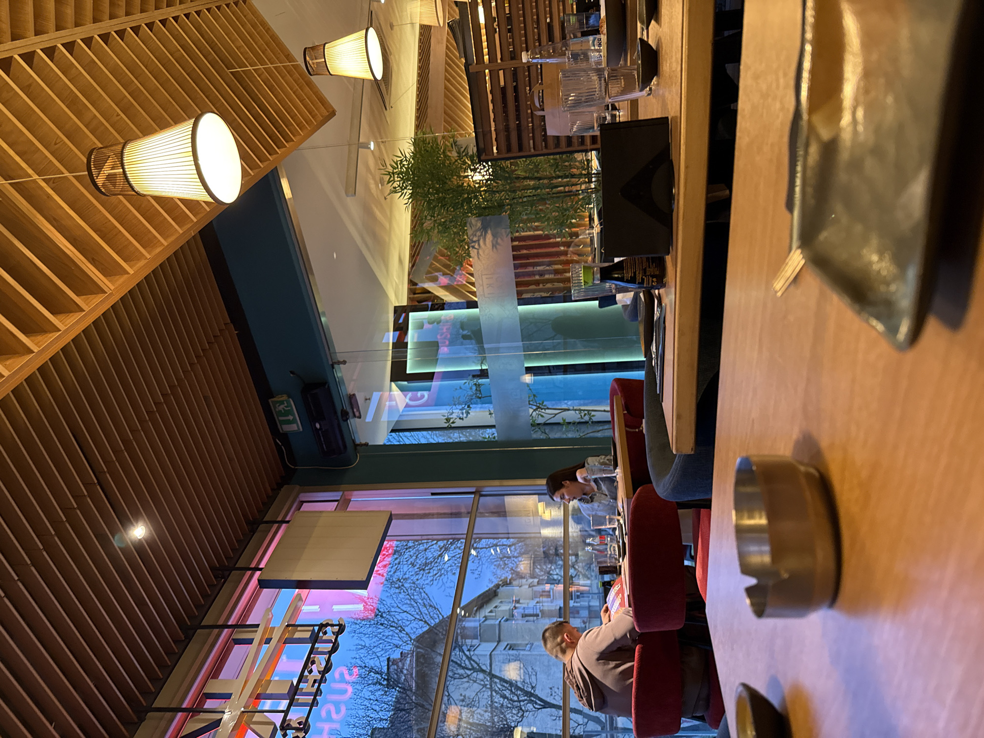 The image shows the interior of a sushi restaurant in Sofia, Bulgaria. The restaurant has large windows looking out onto a street with buildings and bare trees. A "SUSHI" sign with neon lights is visible outside. Two people are seated at a table near the window. One person is looking at a phone. Tables are wooden with tableware set. The ceiling has wooden slats with cylindrical light fixtures. Greenery is placed near a glass partition.