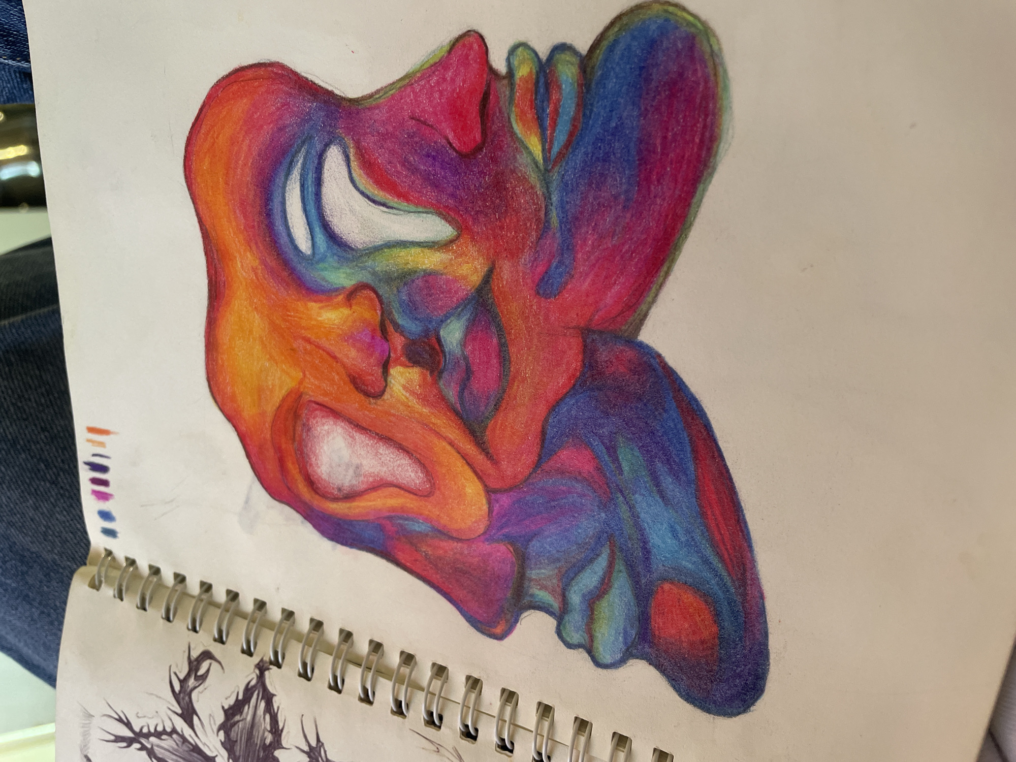 The image shows a colorful pencil drawing in a spiral-bound sketchbook. The drawing appears to depict two faces intertwined, rendered in a vibrant color palette of reds, oranges, yellows, purples, and blues. The faces are abstract and the use of color creates a surreal effect. A set of colored pencil swatches are visible on the top edge of the open sketchbook. The page below the drawing contains another drawing in black and white, and partially visible. The sketchbook rests on a denim-covered surface. There is no information to say the image was taken in Debelt, Bulgaria.