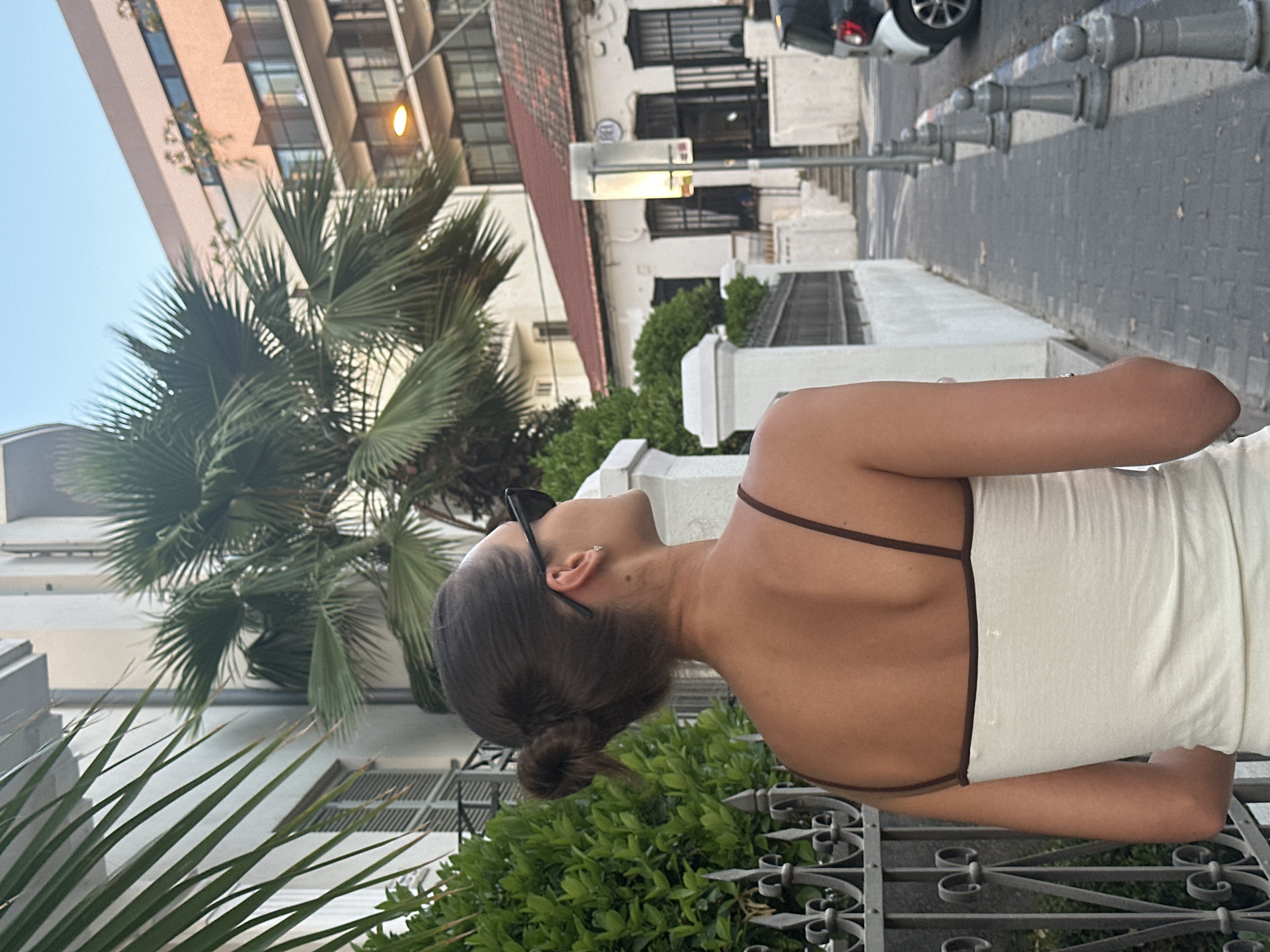 In an urban setting, a young woman with her hair pulled back in a bun is viewed from behind, wearing a white tank top with brown trim and black sunglasses. She is standing on a cobblestone sidewalk next to a decorative metal fence and lush green bushes. In the background, a palm tree stands tall, with buildings of varying architectural styles visible. One building, a white structure with a red-tiled roof, appears to be older, while a more modern, multi-story building with balconies stands behind it. A street lamp illuminates the scene. The woman is looking up and to the right, seemingly observing something in the distance. The image captures a moment on a street in Tel Aviv, Israel, likely during the late afternoon or early evening due to the soft light.