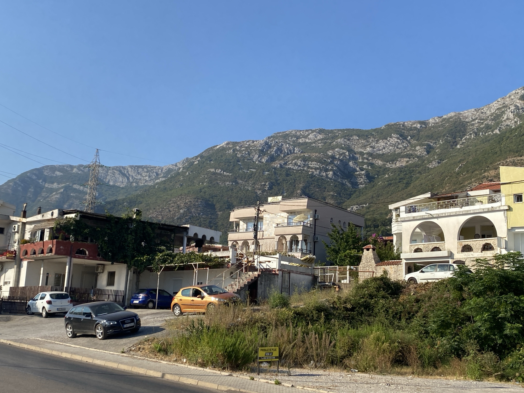 The image shows a residential area in Zgrade, Montenegro, under a clear blue sky. In the foreground, a paved road runs alongside a sidewalk. Beyond the sidewalk is a grassy area with overgrown vegetation. A yellow sign with black text, reading "PRODAJE SE PLAC 636 m² +382 69 013 209", is planted in the grass.
Several cars are parked in front of buildings. A black Audi is parked on the right side of the road, with its front facing towards the left. A white hatchback is parked further back, to the left of the Audi. Behind these cars, a blue sedan is parked in a driveway. An orange hatchback is parked on the right side of the driveway.
The buildings are multi-story structures with balconies and arched windows. The architecture appears to be typical of Mediterranean coastal regions. Some buildings are covered in ivy and other greenery. In the background, a large, rugged mountain range dominates the landscape. A tall electricity pylon is visible on the left side of the image, with power lines stretching across the frame. The overall scene depicts a sunny day in a somewhat rural, mountainous part of a city.