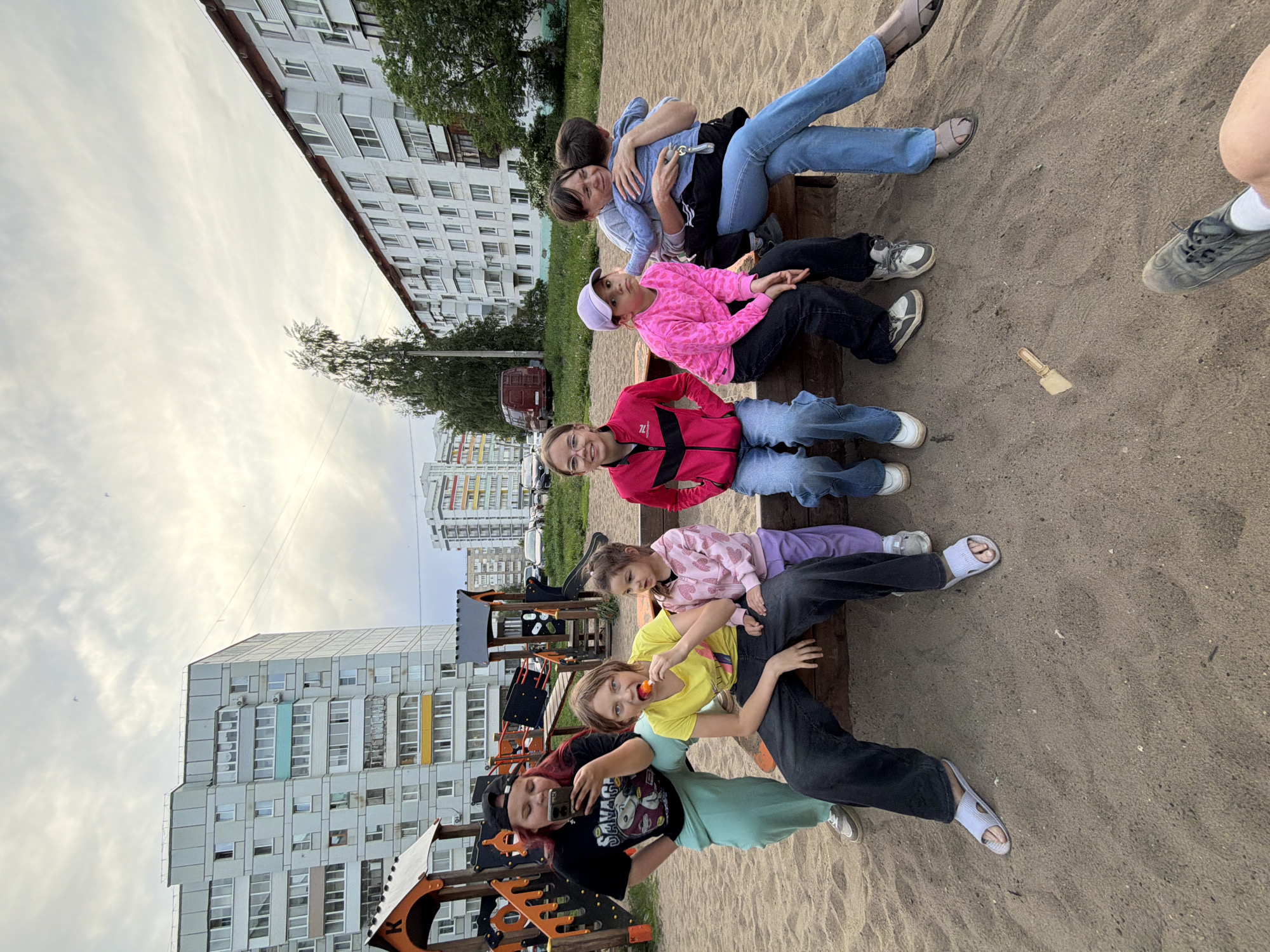 The image captures a group of seven individuals posing on a bench in what appears to be a playground or park setting. The location is identified by the presence of typical urban apartment buildings in the background, a characteristic of Chaikovsky, Russia.
The group consists of a mix of adults and children. They are sitting closely together on the bench, some hugging or leaning on each other, suggesting a familial or friendly relationship. Their attire includes a range of casual clothing such as jeans, jackets, t-shirts, and sneakers or sandals. One individual is holding a mobile phone, possibly taking a selfie or group photo. Another person is holding an orange object.
The environment consists of sandy ground, playground equipment like slides and climbing structures. The apartment buildings are visible in the background, offering a glimpse of urban architecture and daily life. The presence of trees and grass indicates a mix of urban and natural elements. The sky is overcast or cloudy.