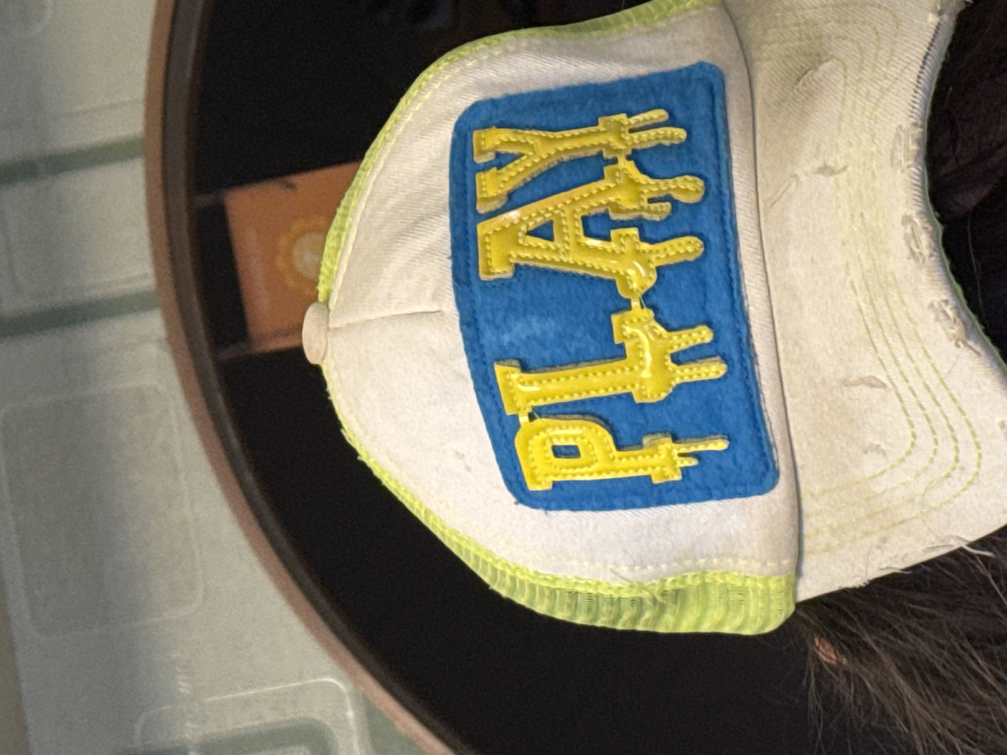A close-up shot shows a partially visible white baseball cap resting on a dark surface. The cap features a blue rectangular patch with the word "PLAY" embroidered in yellow, vertically oriented. A thin band of green piping is visible along the brim's edge. The cap's fabric appears worn and slightly discolored. A small portion of dark hair is visible near the cap's edge. To the left, a portion of a brown shelf and a lighter-colored, textured surface are visible. There is no indication of a location in Novo-Nikol'skoe, Russia.