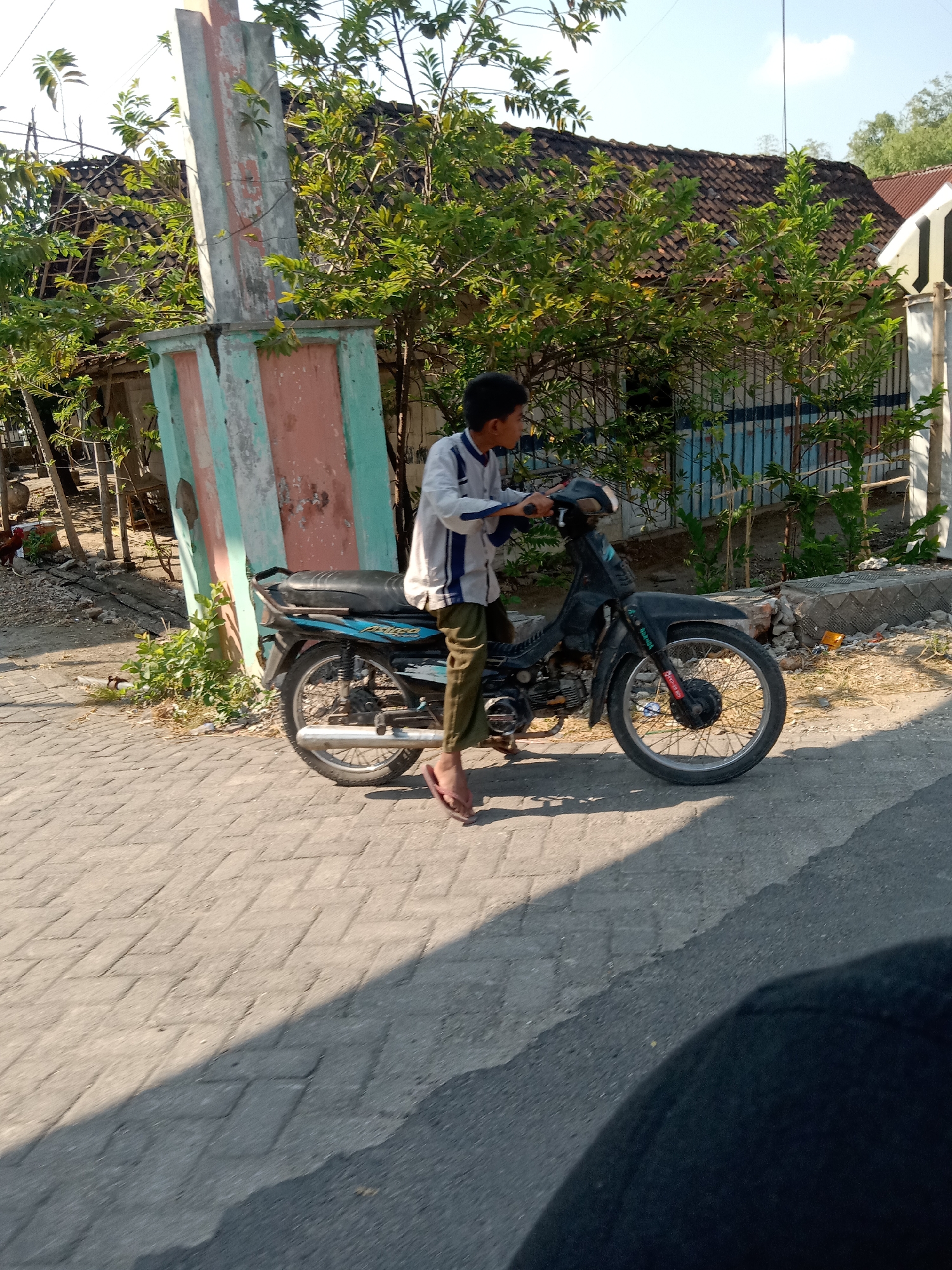 A young boy, appearing to be of Indonesian descent and wearing a white and blue striped long-sleeved shirt, olive-green pants, and pink flip-flops, is pushing a dark-colored motorcycle on a paved brick sidewalk in Semawot, Indonesia. The motorcycle has a black seat and blue accents. The boy is looking towards the right of the frame. Behind him, a weathered, multi-colored pillar with vertical stripes of peach, light green, and white stands prominently. Lush green trees and foliage surround the pillar and extend to the background, where a house with a dark-tiled roof is visible. A fence with vertical blue bars is also present in the background. The sun is casting a strong shadow across the sidewalk from the left side of the frame. The overall environment suggests a sunny day in a rural or semi-urban area.
