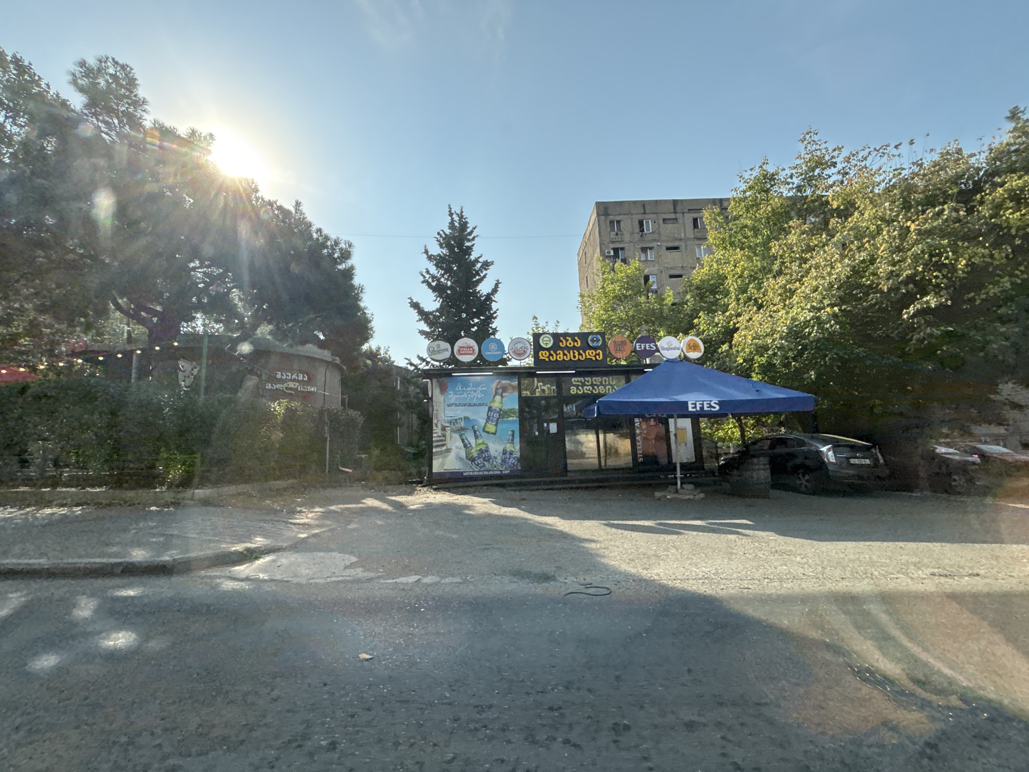 The image captures a street-level view of a small commercial establishment with an outdoor seating area, likely a cafe or bar, located in Tbilisi, Georgia. The sun is shining brightly, casting lens flares and dappling the asphalt with light. In the foreground, the asphalt road shows some wear and tear, with a curb bordering a dirt or gravel area.
The establishment itself appears to be a kiosk-like structure with a blue awning that has "EFES" written on it. Behind the awning, there are windows and doors. A large advertisement featuring bottles of Efes beer is displayed on the side of the structure. Several round signs with logos of different beer brands, such as Stella Artois, are mounted on poles above the establishment.
To the left of the establishment, there are trees and bushes, with some strings of lights suggesting an outdoor seating area. To the right, a dark-colored car is parked near the establishment, partially obscured by the awning and some barrels. Further to the right, a part of a red car is visible. In the background, a multi-story concrete apartment building stands, characteristic of Soviet-era architecture, with several visible windows. Lush green trees surround the area, and a tall, slender evergreen tree stands prominently behind the establishment. The sky is clear and blue.
