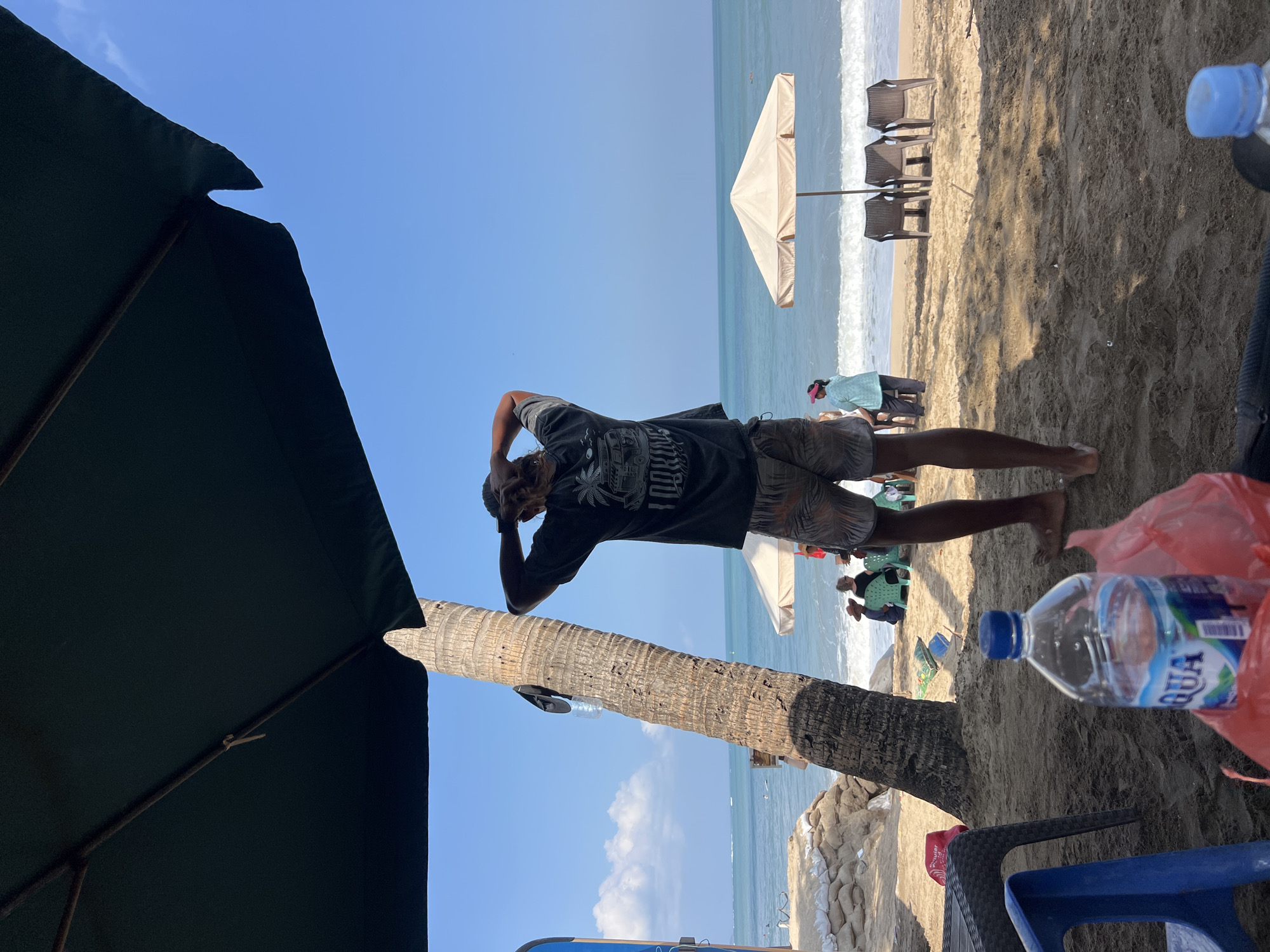 A man with long hair stands on a sandy beach in Kuta, Indonesia, with his back to the camera, touching his hair. He is wearing a dark t-shirt with a graphic print and patterned shorts. In the foreground, a bottle of Aqua water and a red plastic bag are visible. Beyond the man, the ocean meets the shore with small waves. Beach umbrellas and chairs are set up on the sand. In the distance, other beachgoers are scattered, and sandbags are piled along the coastline. The sky is clear blue with a few scattered clouds.