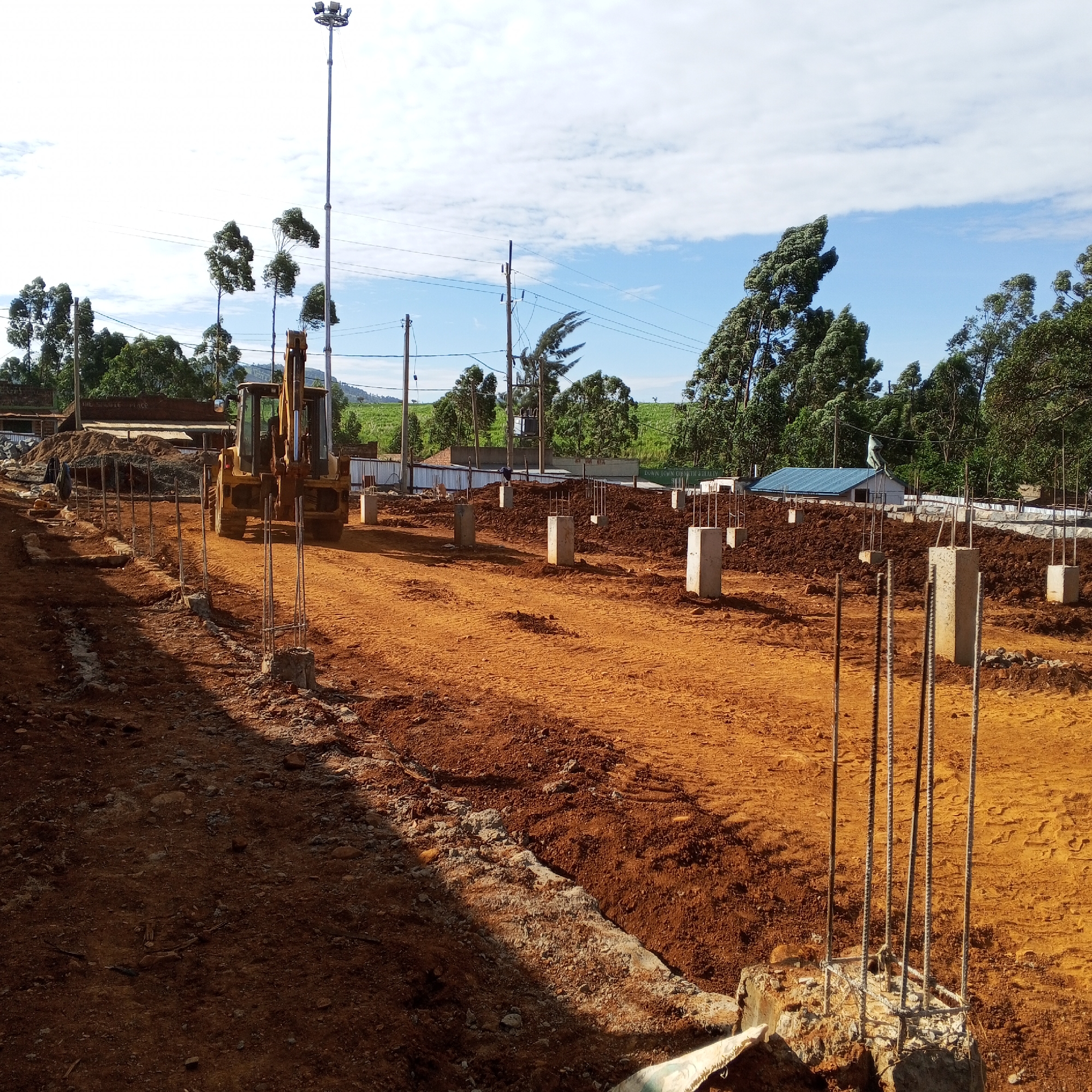 The image depicts a construction site in Borabu Chitago ward, Kenya. The ground is red earth, partially leveled. Several concrete pillars with rebar extending upwards are erected in a grid pattern. A yellow backhoe is parked on the left side of the frame. In the background, trees and buildings are visible, along with utility poles and wires. The sky is partly cloudy.
