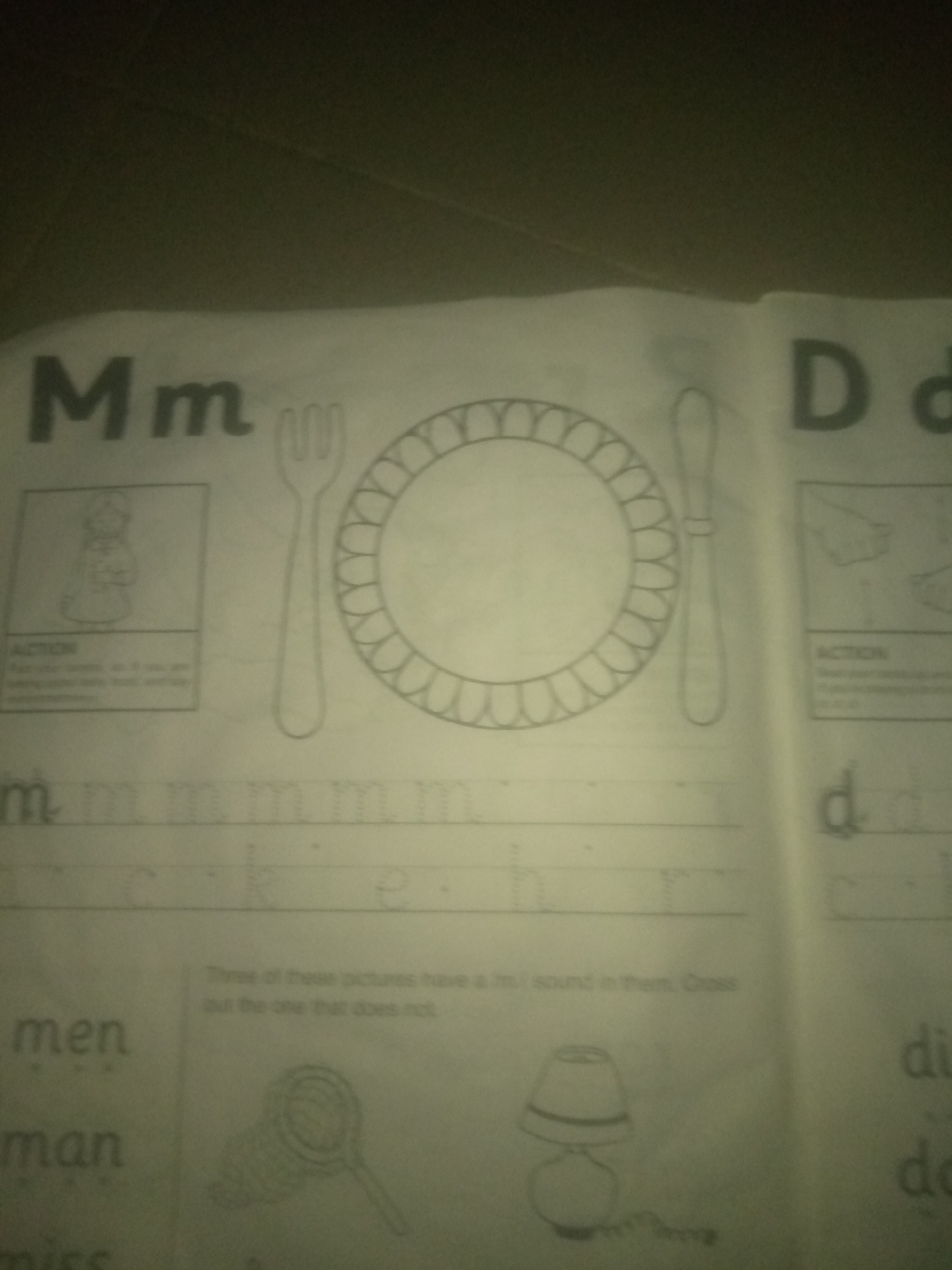 The image is a close-up, slightly angled shot of a page from what appears to be a children's educational workbook. The page is predominantly white with black print and line drawings.
On the left side of the page, the letters "Mm" are prominently displayed in large font. Below this, a framed image shows a person in traditional attire. Text, likely instructions, is present below this frame and is partially obscured. Further down, the letter "m" is traced multiple times on lines, indicating a handwriting practice exercise. To the left of these traced letters, the words "men", "man", and "miss" are printed.
The center of the page features a large outline drawing of a plate, a fork, and a knife. Below this, a line of text reads, "Three of these pictures have a /m/ sound in them. Cross out the one that does not."
On the right side of the page, the letters "D d" are visible in large font, similar to the "Mm" on the left. Below this, a framed image depicts hands. Text labeled "ACTION" is present below this frame. Further down, the letters "d" and "di" are visible, suggesting another section for letter practice.
The lower portion of the page shows line drawings of a magnifying glass and a lamp. The overall impression is of a lesson focusing on letters and sounds, likely for young learners. There is no discernible connection to the city of Jauro Yinu, Nigeria, as the image depicts a generic educational worksheet. The lighting suggests the photo was taken in an indoor environment with artificial light, possibly a flash. The page appears to be lying flat on a surface, and the background is dark and out of focus.