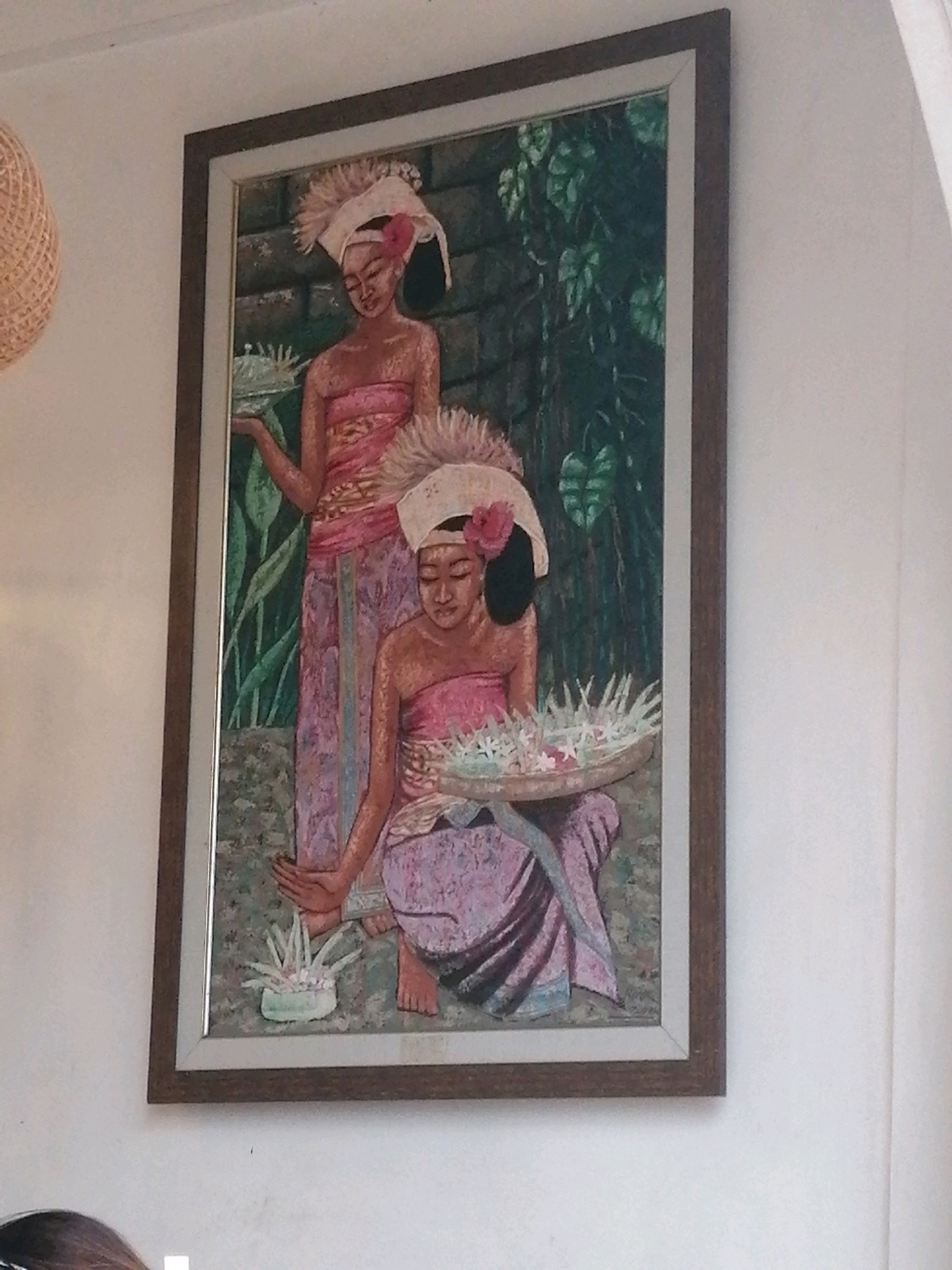 A framed painting depicts two young women in traditional Indonesian attire, likely from Ubud, Indonesia. The woman in the foreground is kneeling and holding a woven tray filled with flowers. She wears a pink, strapless top and a purple sarong. Her hair is adorned with a white headdress topped with a pink flower. The woman behind her stands with her arms outstretched, wearing a similar pink top and purple sarong, and a more elaborate white headdress. Both women have dark hair and fair skin. The background of the painting features a stone wall and lush green foliage. The painting is framed in a wooden border and hangs on a white wall. To the left of the painting, a portion of a woven circular object is visible.
