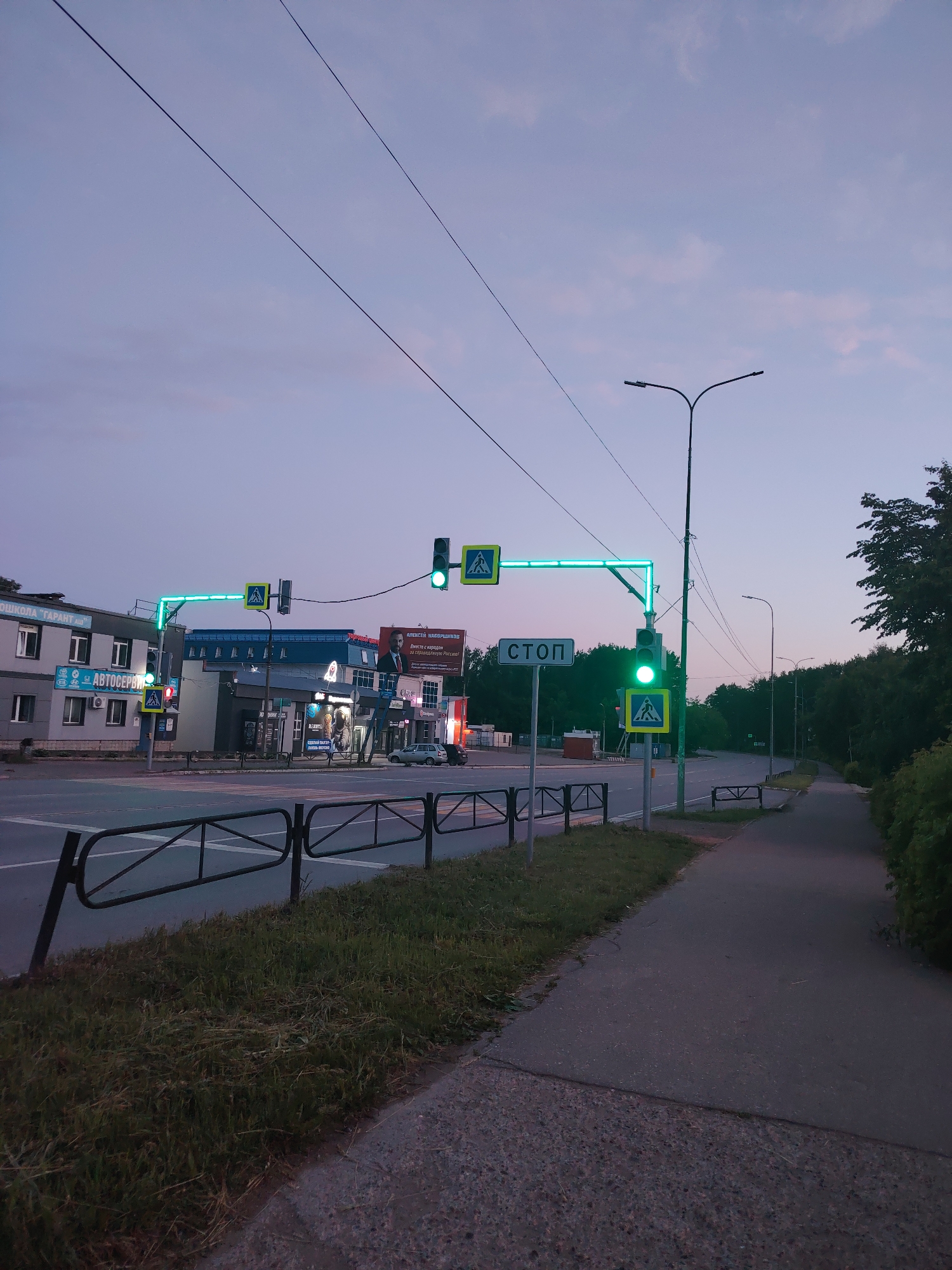 This media file captures a quiet urban street scene during dusk or early night in Chaikovsky, Russia. The sky, a mix of soft purples and pinks, indicates the twilight hours, with overhead power lines traversing the frame.
The foreground features a paved sidewalk on the right and a patch of grass with a metal pedestrian fence on the left, separating the sidewalk from a multi-lane road. The road itself is visible, with lane markings faded but discernible.
In the midground, a prominent set of traffic lights is visible, with the main light illuminated green, indicating an open flow of traffic. Another traffic light further down the road also shows green. A large, bright green illuminated structure outlines the pedestrian crossing area above the road. Several yellow and black pedestrian crossing signs are mounted near the traffic lights. A white "СТОП" (STOP) sign on a pole is clearly visible to the right of the main traffic light. Streetlights are present but not fully illuminating the scene, complementing the natural twilight.
On the left side of the road, several low-rise commercial buildings line the street. Visible text on these buildings includes "ШКОЛА 'ГАРАНТ АВТО'" (GARANT AUTO SCHOOL) and "АВТОСЕРВИС" (CAR SERVICE). Further down, a large billboard displays a picture of a man (likely Alexei Navalny, with "АЛЕКСЕЙ НАВАЛЬНЫЙ" visible) and Russian text that appears to be political or commercial in nature. A few parked cars can be seen near these buildings.
The right side of the image shows a tree-lined area extending into the distance, suggesting a park or forested edge alongside the road. The overall impression is one of a calm, transitioning period between day and night in a functional urban environment.