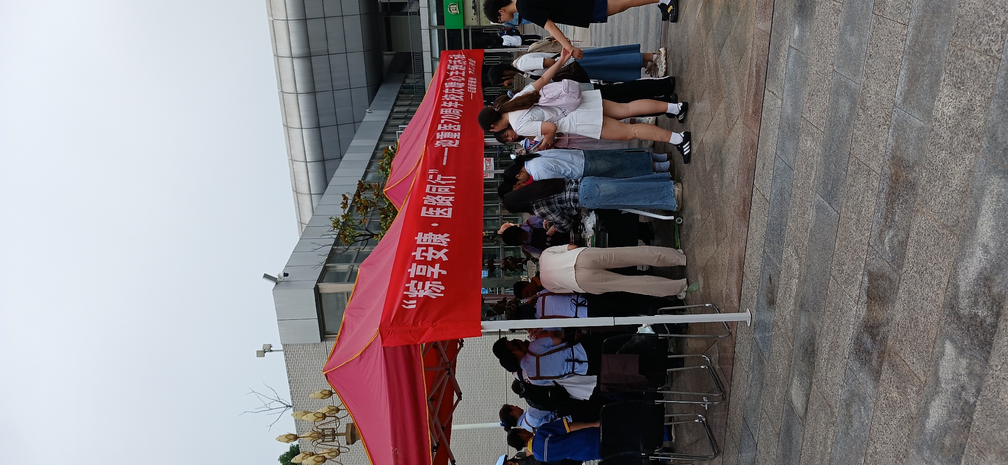 A group of people are gathered around a red tent, likely at an outdoor event. The tent has white text printed on it in Chinese characters, which appears to be a slogan or title. Some individuals are standing and observing, while others are interacting with items on a table beneath the tent. One person in a light-colored shirt and pants is bent over the table, possibly engaged in an activity or distribution. Several people are wearing casual clothing, and some are in what might be uniforms or matching attire. The ground is paved with stone tiles, and there are signs of recent rain with some areas appearing wet. In the background, there is a modern building with a metallic facade and windows. The overall scene suggests an organized public gathering or a promotional event.