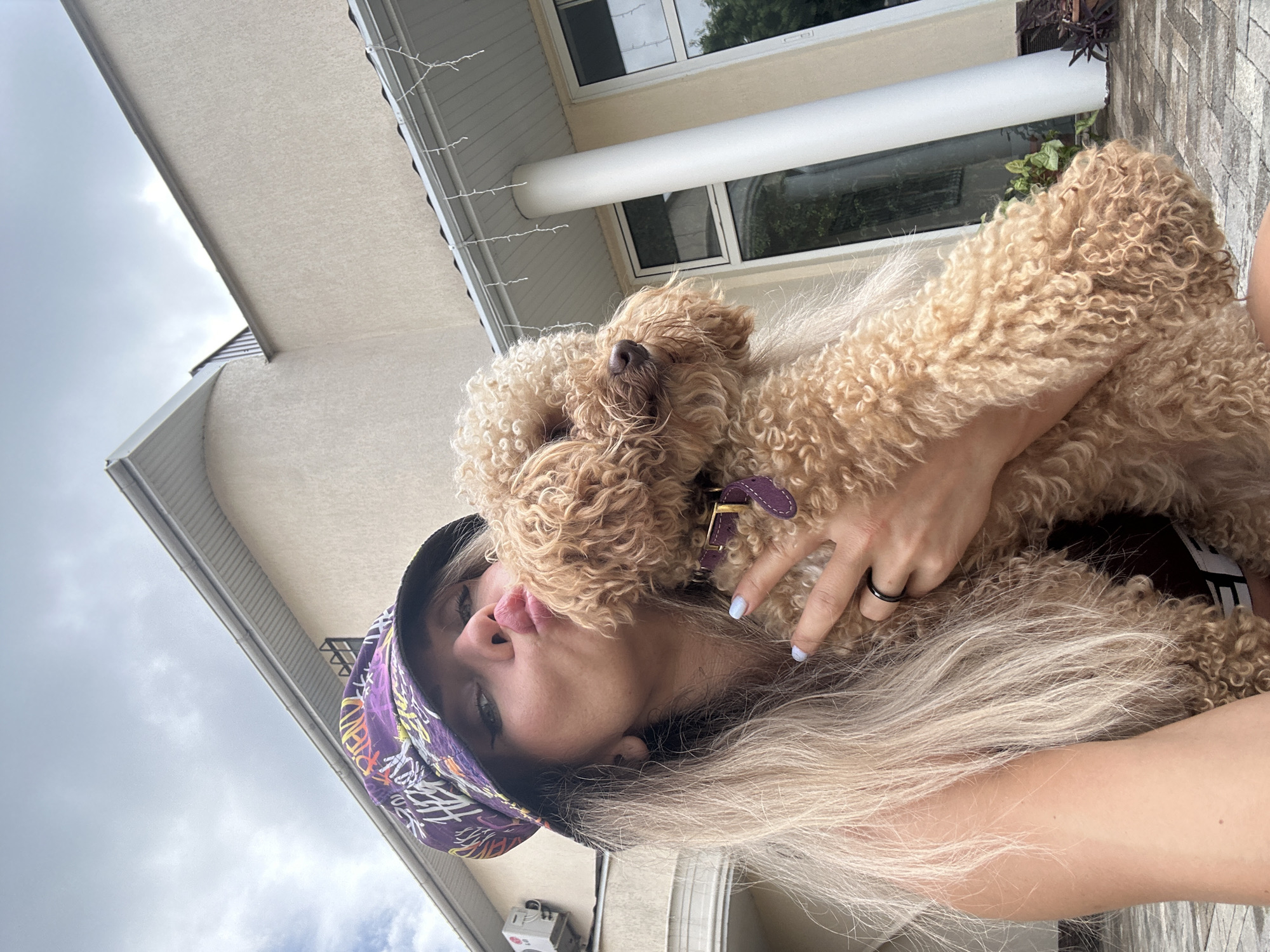 A blonde woman wearing a purple bucket hat with graffiti print and a black tank top is taking a selfie with a light brown poodle in Armavir, Russia. The woman is puckering her lips towards the dog, who is looking off to the side. Her left hand is around the dog's neck, and her right hand is holding the dog up, with a black ring on her ring finger and light blue nail polish on her fingernails. The background shows a tan, stucco-sided house with white columns, a white gutter, and string lights. The sky is cloudy.