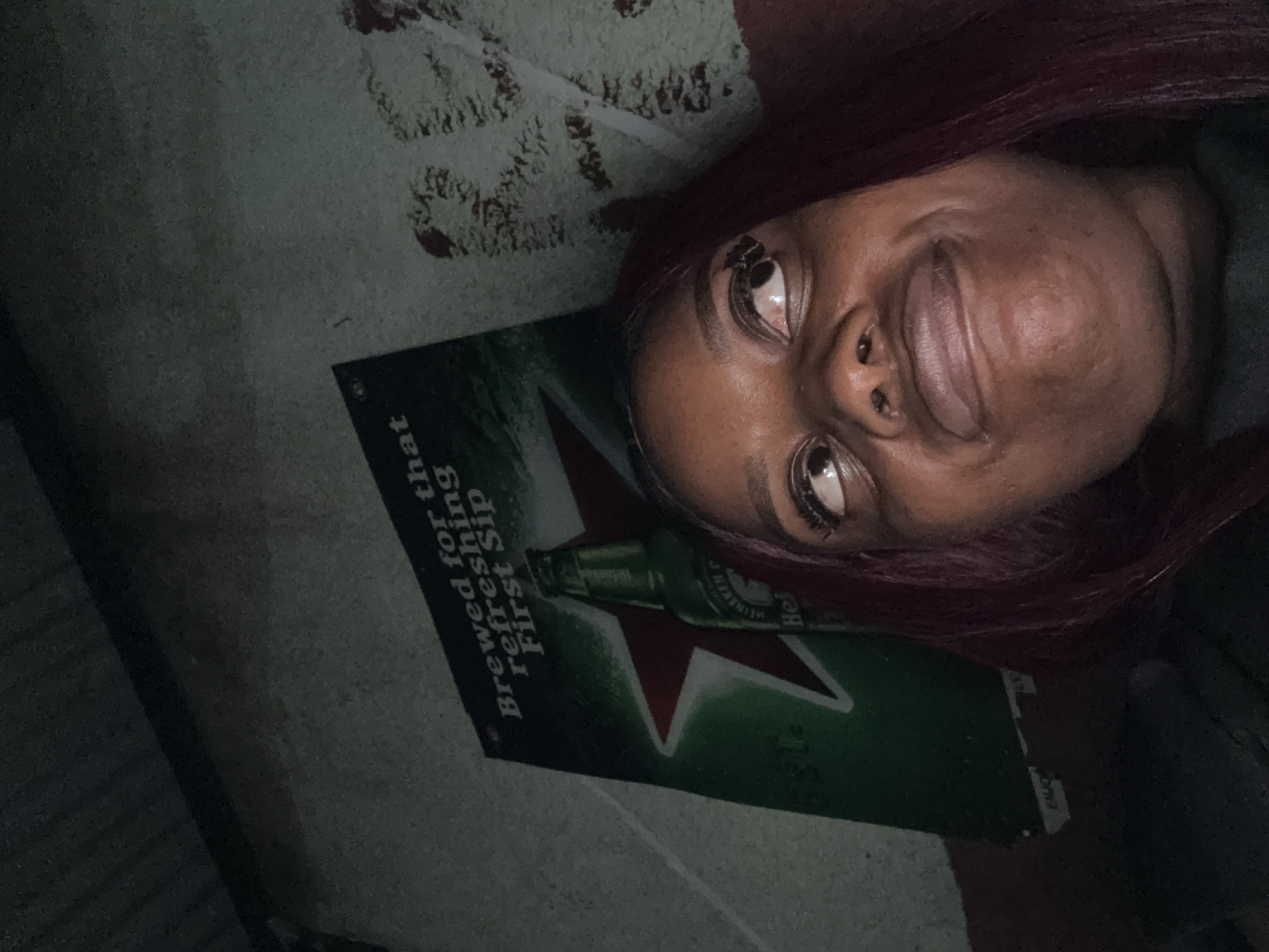 The image shows a close-up of a young Black woman with purple hair looking upwards with a playful expression. Her eyes are wide and her mouth is slightly open. In the background, a Heineken beer poster is visible, featuring a green bottle and a red star. The poster appears to be affixed to a textured, light-colored wall. The lighting is dim, casting shadows and creating a somewhat moody atmosphere. The scene appears to be indoors, possibly in a casual setting. There are no visible indicators of the specific location within Abuja, Nigeria, beyond the poster itself which is a global brand.