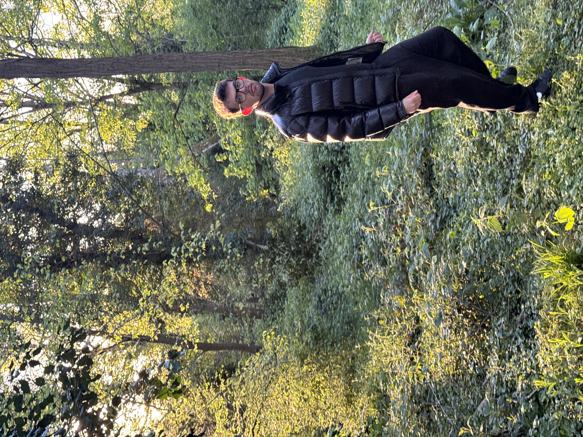 The image, oriented correctly, depicts a man standing outdoors in a natural, wooded setting. The subject is a young to middle-aged man, with short dark hair, wearing glasses, a black puffer jacket, and black trousers. He is looking directly at the camera with a neutral expression.
The scene is dominated by dense green foliage and trees, suggesting a forest, park, or natural reserve. The ground appears to be covered in undergrowth and low-lying plants. Several tree trunks are visible in the background, with their branches reaching upwards, adorned with light green leaves.
The time of day appears to be late afternoon or early evening, characterized by the warm, golden light of the sun filtering through the tree canopy from behind the man. This backlighting creates a bright halo effect around his head and shoulders, particularly on his right ear and the rim of his glasses, and illuminates the leaves with a vibrant, glowing quality. The light also casts bright patches on the surrounding vegetation.
There is no specific activity taking place other than the man posing for the photo. The overall atmosphere is tranquil, captured during a sunny period. The location context provided indicates this scene is in Bures-sur-Yvette, France. No visible text is present in the image.
