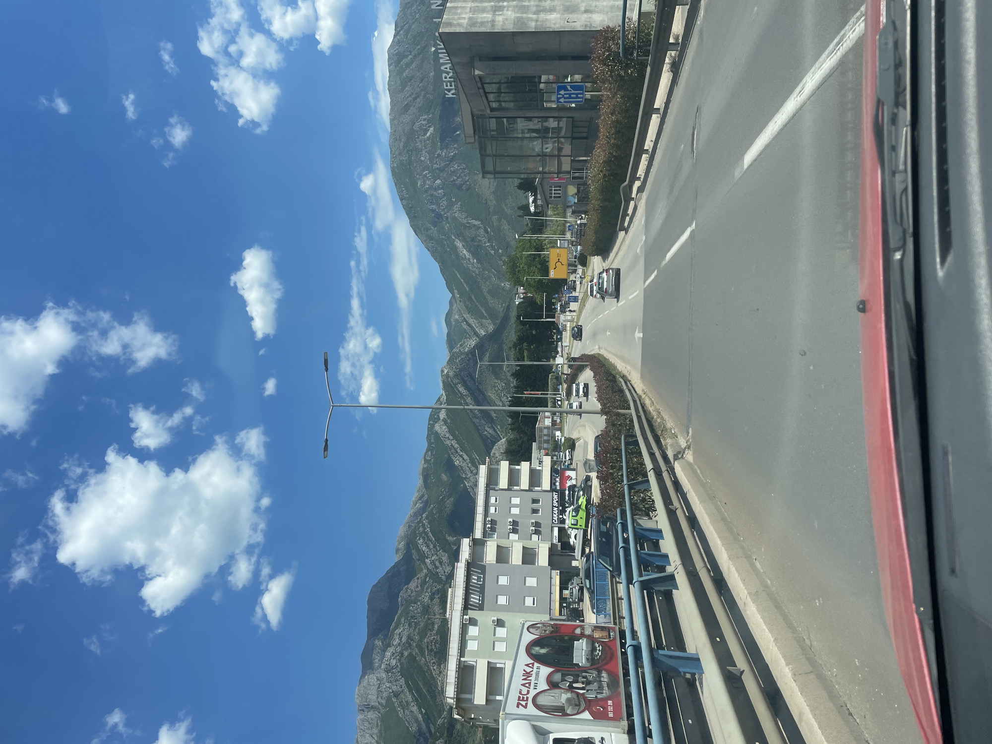 The image depicts a road in Bjeliši, Montenegro, as seen from a moving vehicle. A mountain range dominates the background under a blue sky with scattered clouds. A building with the word "KERAMIKA" visible on its facade stands on the right side of the road. Cars and street lights line the street, and a bridge with a blue railing is visible in the foreground. The side of a white truck, bearing the logo of "ZECANKA," is also present.