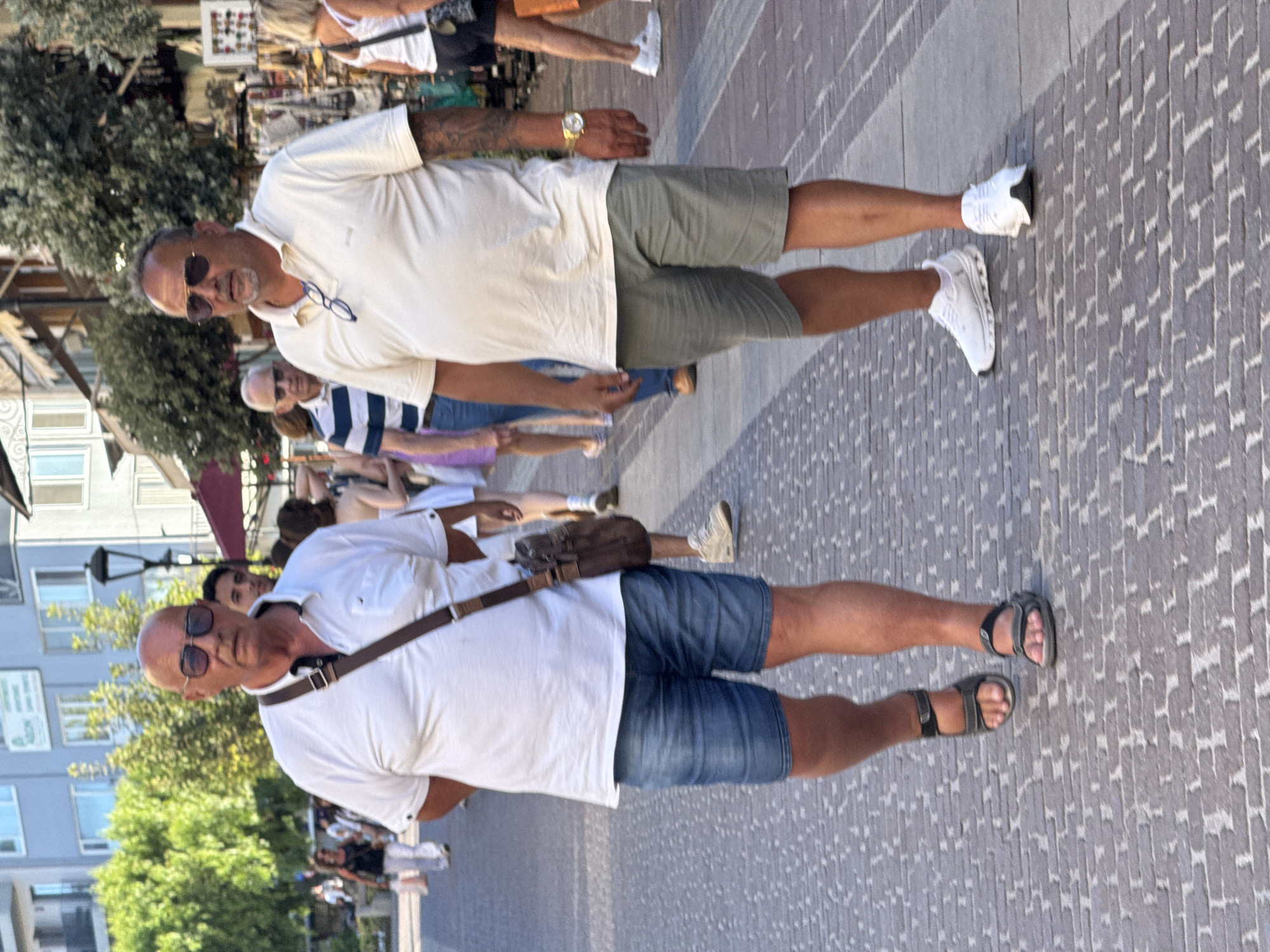 Two adult males are walking on a cobblestone pedestrian street in Chania, Greece. The man on the left is wearing a white polo shirt, blue denim shorts, and dark sandals. He has a brown satchel slung across his body and is wearing sunglasses. The man on the right is wearing a light-colored polo shirt, green shorts, and white athletic shoes. He has sunglasses resting on his shirt collar and is wearing a gold wristwatch. Several other pedestrians are visible in the background, walking in the same direction. Buildings line the street, with shops and trees on either side. The lighting suggests it is daytime, possibly late afternoon, with the sun casting long shadows.