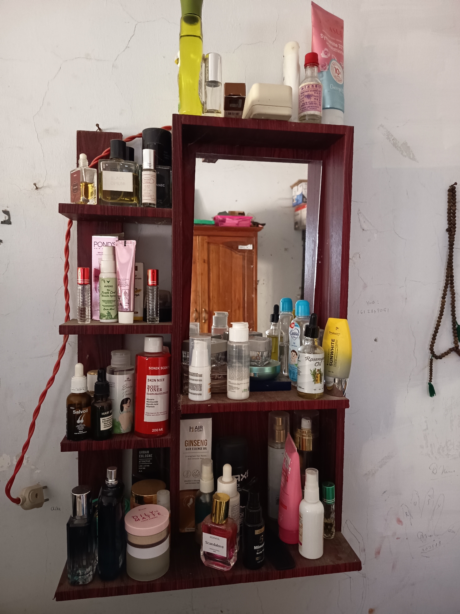 The image depicts a shelf unit containing various cosmetic and personal care products. The shelf unit is made of dark wood and has a mirror integrated into its design. It is mounted on a cracked, off-white wall. The products on the shelf include bottles of perfume, toners, mouth spray, and lotions. Some of the visible brands are POND'S, SALVOIL, and SUNWHITE. There are also several unidentifiable bottles and containers. A beaded necklace with green tassels hangs on the right side of the wall. The overall scene suggests a personal grooming area, likely a vanity or dressing area. The presence of graffiti on the wall indicates the location is likely within a residential or public space.
