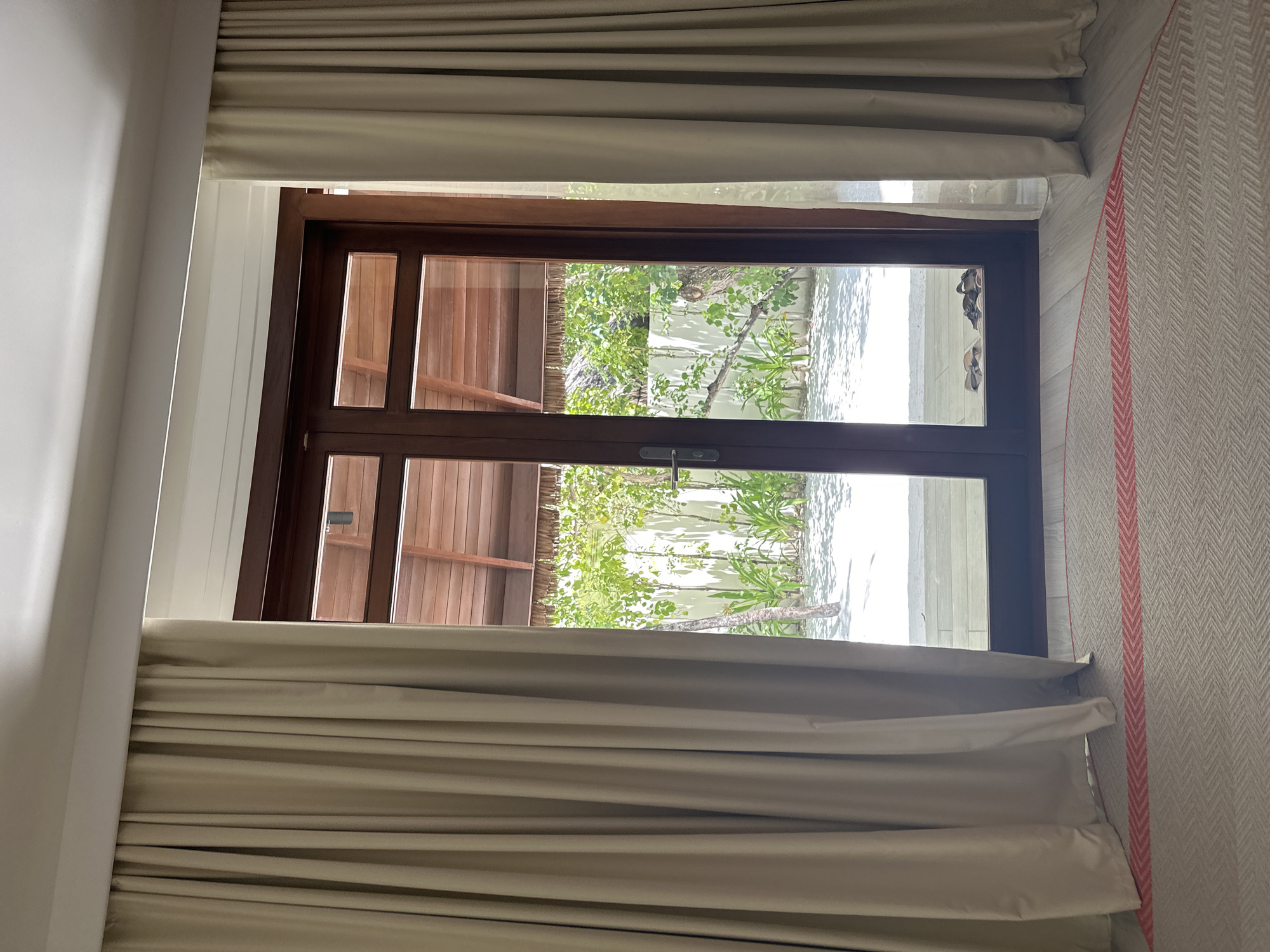 This image shows a view from inside a room looking out through a set of double glass doors to a tropical outdoor area, likely in Halaveli, Maldives. The doors are framed in dark wood and are partially open, with sheer white curtains drawn back on either side. Through the glass, a sandy courtyard with lush green foliage and trees is visible. The architecture of the outdoor area features a thatched roof structure. Inside the room, a portion of a rug with a chevron pattern and a red stripe is visible on the floor. A pair of sandals rests just outside the doors on the sandy ground. The overall scene suggests a tranquil and luxurious resort setting.