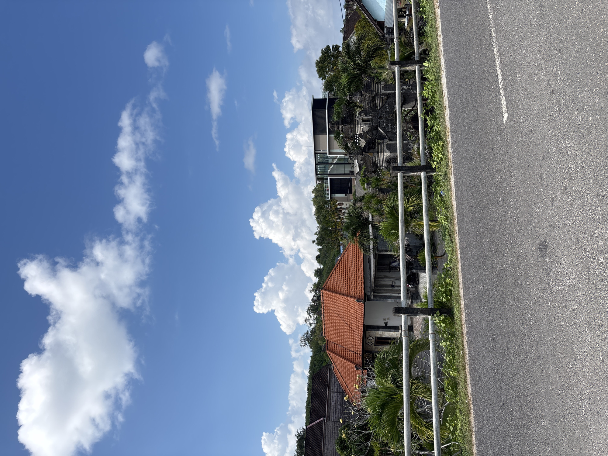 The image captures a bright, sunny daytime scene along a road in Kuta Selatan, Indonesia. On the right side of the frame, an asphalt road with a single white line runs vertically, indicating a paved street. A metal railing, consisting of grey vertical posts and horizontal bars, separates the road from the adjacent properties.
Behind the railing, a strip of green grass and foliage is visible, followed by several buildings nestled among lush tropical vegetation, including numerous palm trees. One prominent building features a distinctive reddish-orange tiled roof, suggesting traditional architecture, while another building further back appears more modern with dark grey and glass elements. The overall setting implies a residential or resort area.
The sky is a clear, vibrant blue, dotted with scattered white cumulus clouds of varying sizes and shapes. The bright sunlight casts clear shadows, confirming it is daytime, likely midday or early afternoon. No people or visible text are present in the image, and no specific activity or event is taking place, presenting a tranquil roadside view.