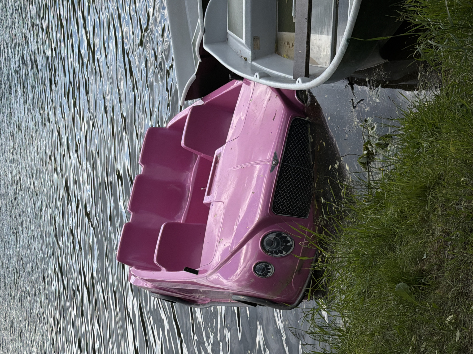 A pink pedal boat, designed to resemble a car front, is partially submerged in a body of water with visible ripples. The boat has seats and a front grille, suggesting a car-like design. Adjacent to the boat and the water's edge is a patch of dense green grass. Another boat, possibly a larger one, is visible at the top of the frame. The location is Malakhovka, Russia.