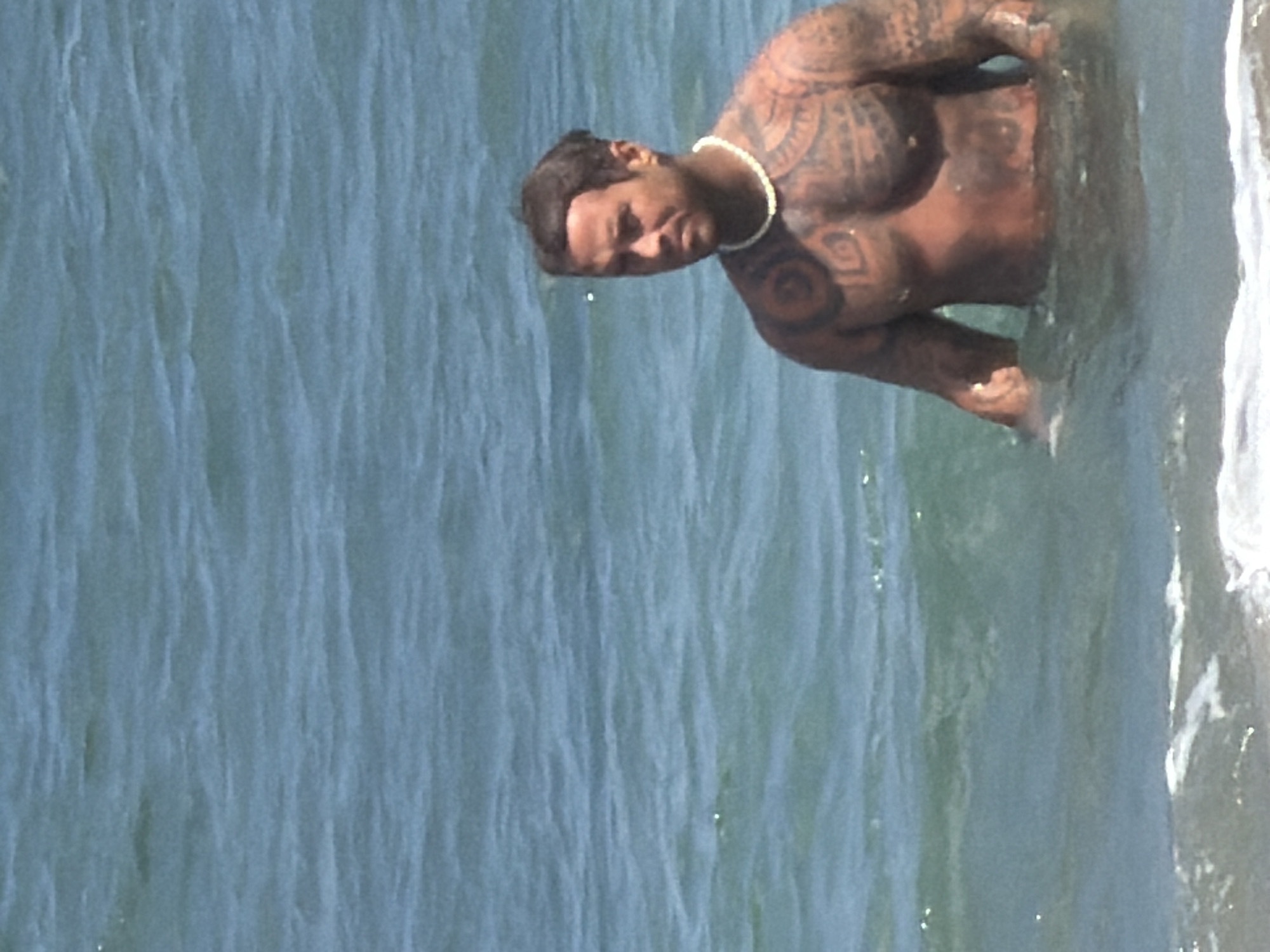 A man with extensive tribal tattoos covering his chest and arms stands waist-deep in the ocean. He is wearing a white beaded necklace. The water is blue and slightly choppy, with small waves breaking near the shore. The background is out of focus, showing more of the ocean under a bright sky. The scene appears to be a tropical beach in North Kuta, Indonesia.